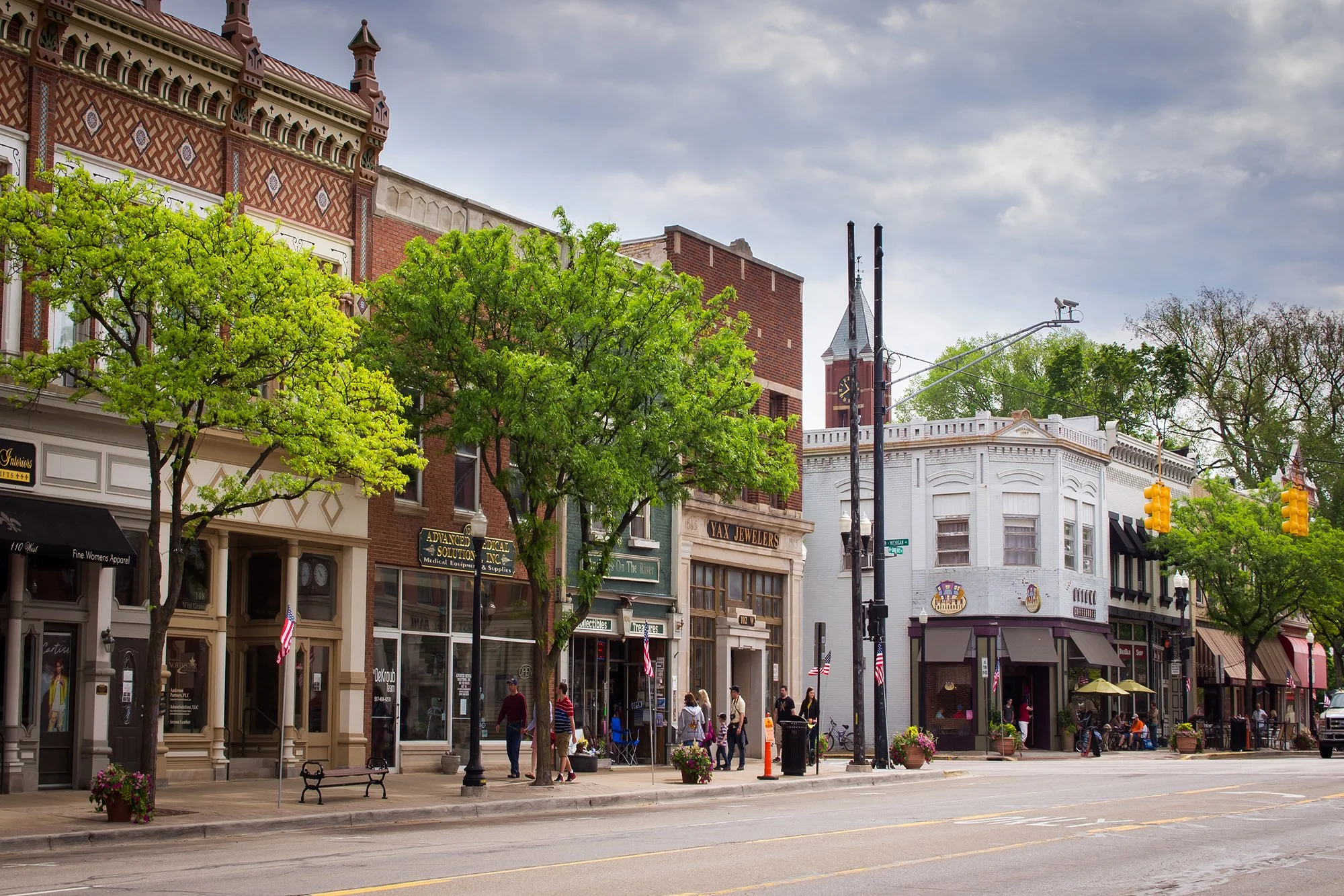 Downtown main street with historic buildings, trees, pedestrians, and outdoor cafes under cloudy sky.