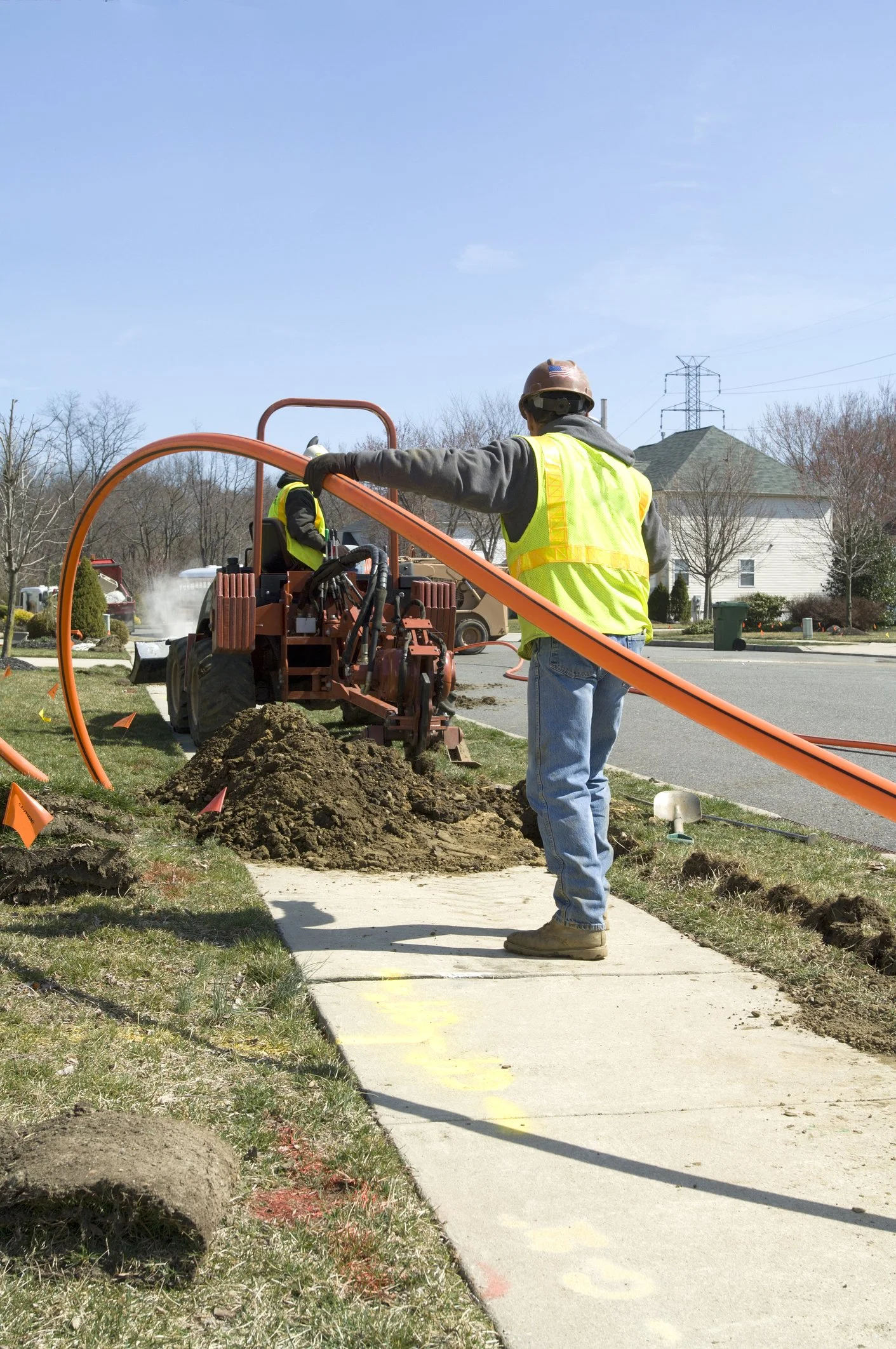 Construction workers installing a sidewalk with equipment and dirt on the grass and sidewalk, residential houses and trees in the background.