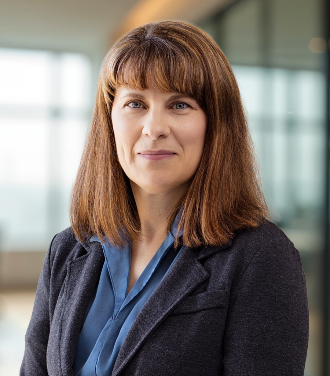 A woman with shoulder-length brown hair and blue eyes, wearing a dark blazer and blue blouse, standing in an office environment.
