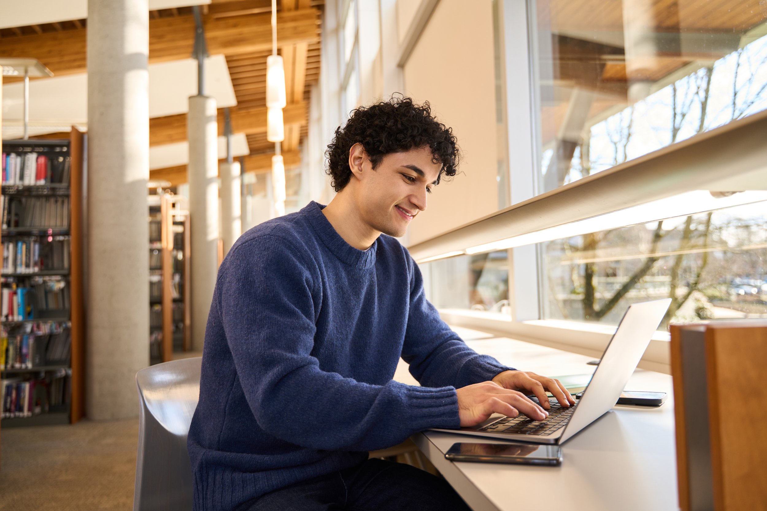 A young man with curly dark hair wearing a navy sweater, sitting at a white table in a modern library, smiling while working on a laptop.
