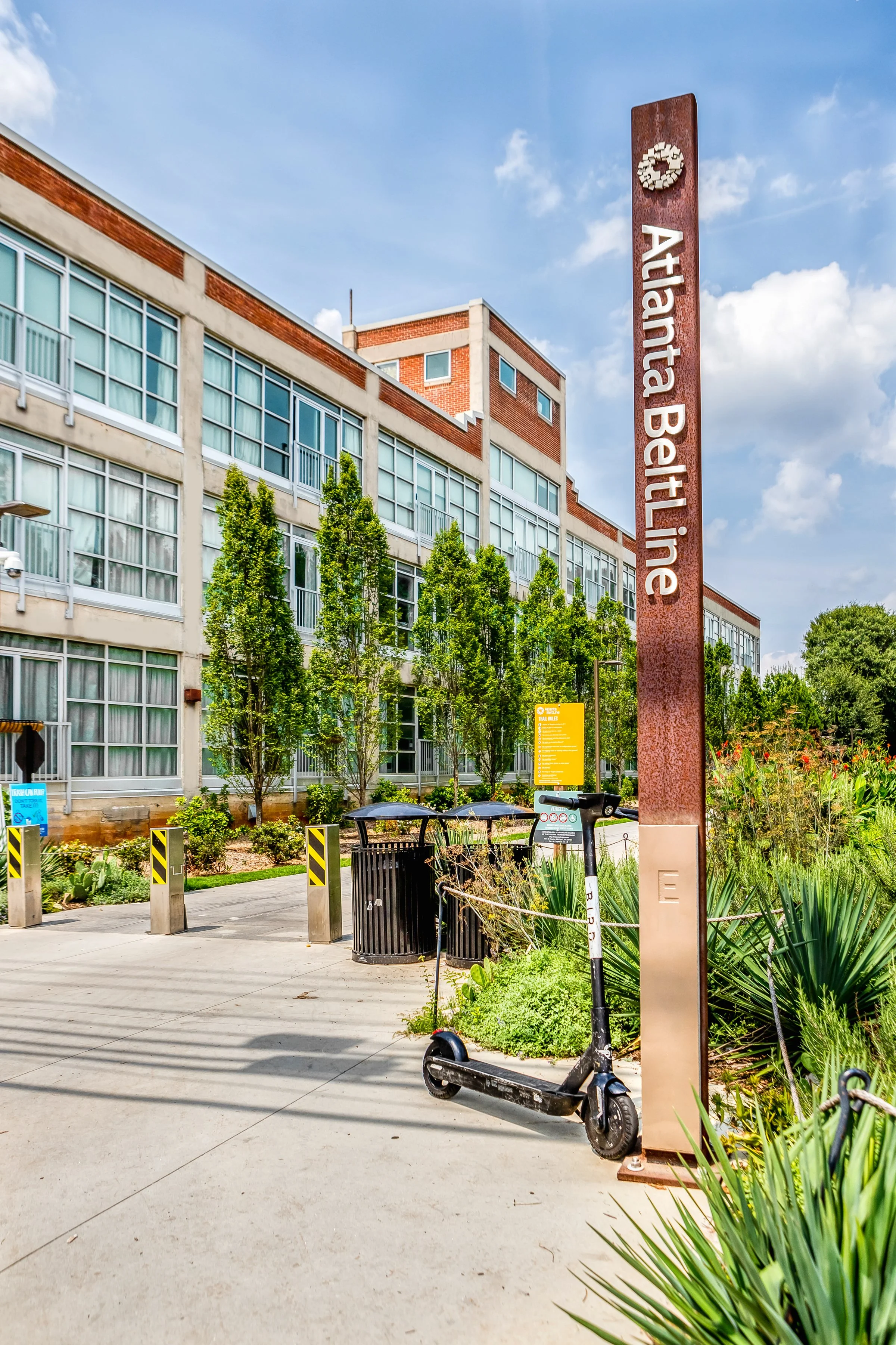 The image shows a sunny urban street scene outside Atlanta Beltline with a tall rust-colored sign with white text, trees, a scooter, a trash can, and a modern building with large windows under a partly cloudy sky.