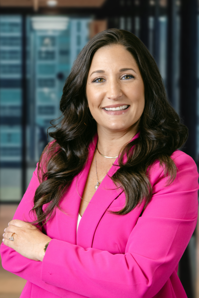 A smiling woman with long dark hair wearing a pink blazer in an office setting.