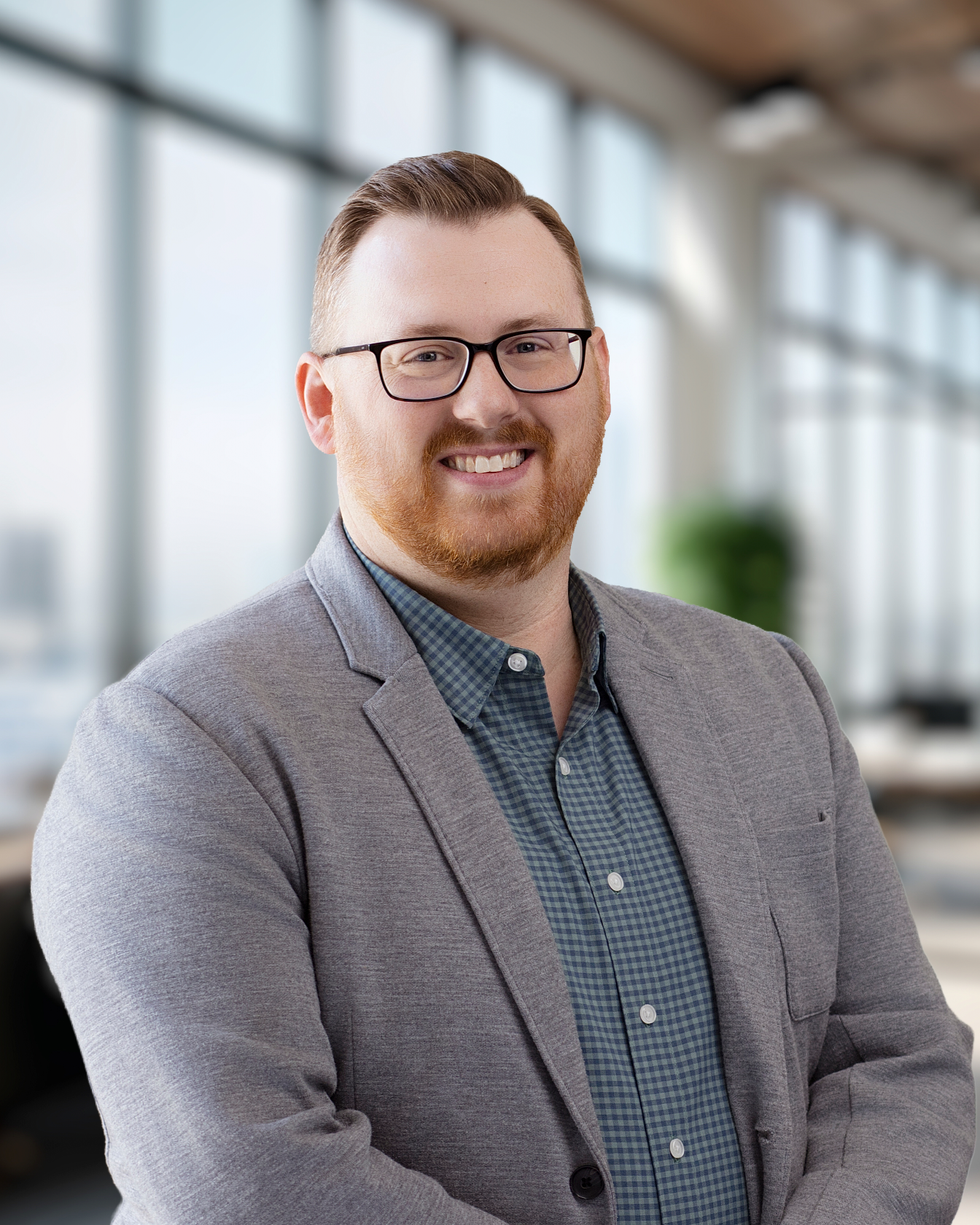 A smiling man with glasses, red hair, and a beard wearing a gray blazer and a blue checkered shirt standing in an office with large windows.