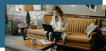 Woman sitting on a mustard-colored sofa working on a laptop in a modern living room with brick wall and decor.