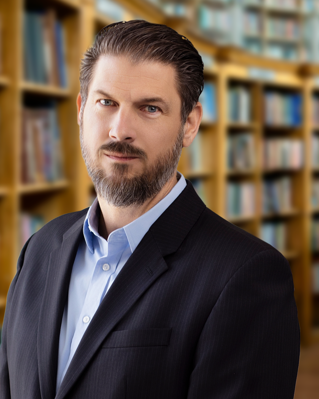 Portrait of a middle-aged man with a beard and slicked-back hair, wearing a dark suit and light blue shirt, in a library with wooden bookshelves filled with books in the background.