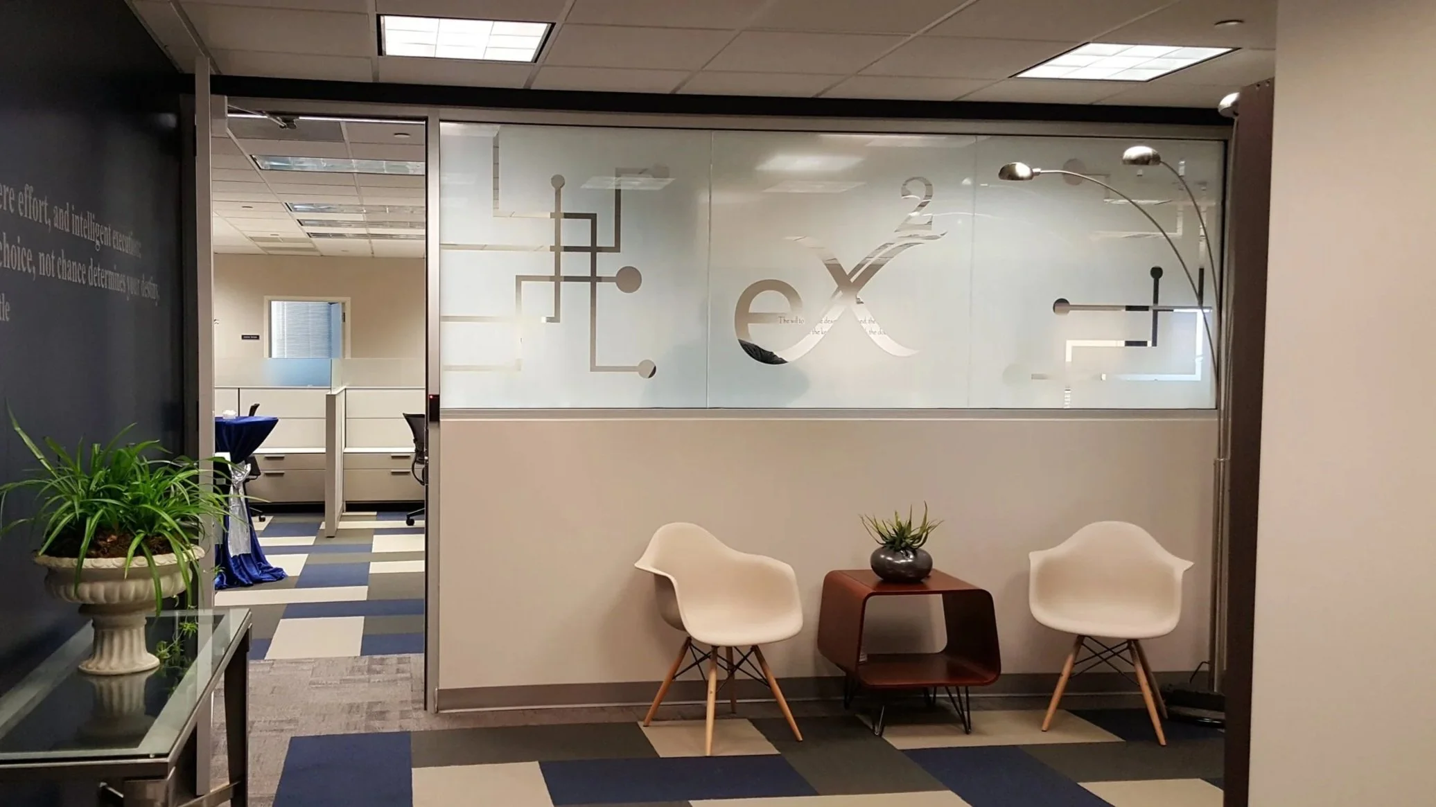 Office corridor with glass walls featuring a logo, two white chairs and a small table with a plant in the foreground, and another office space with desks and chairs in the background.
