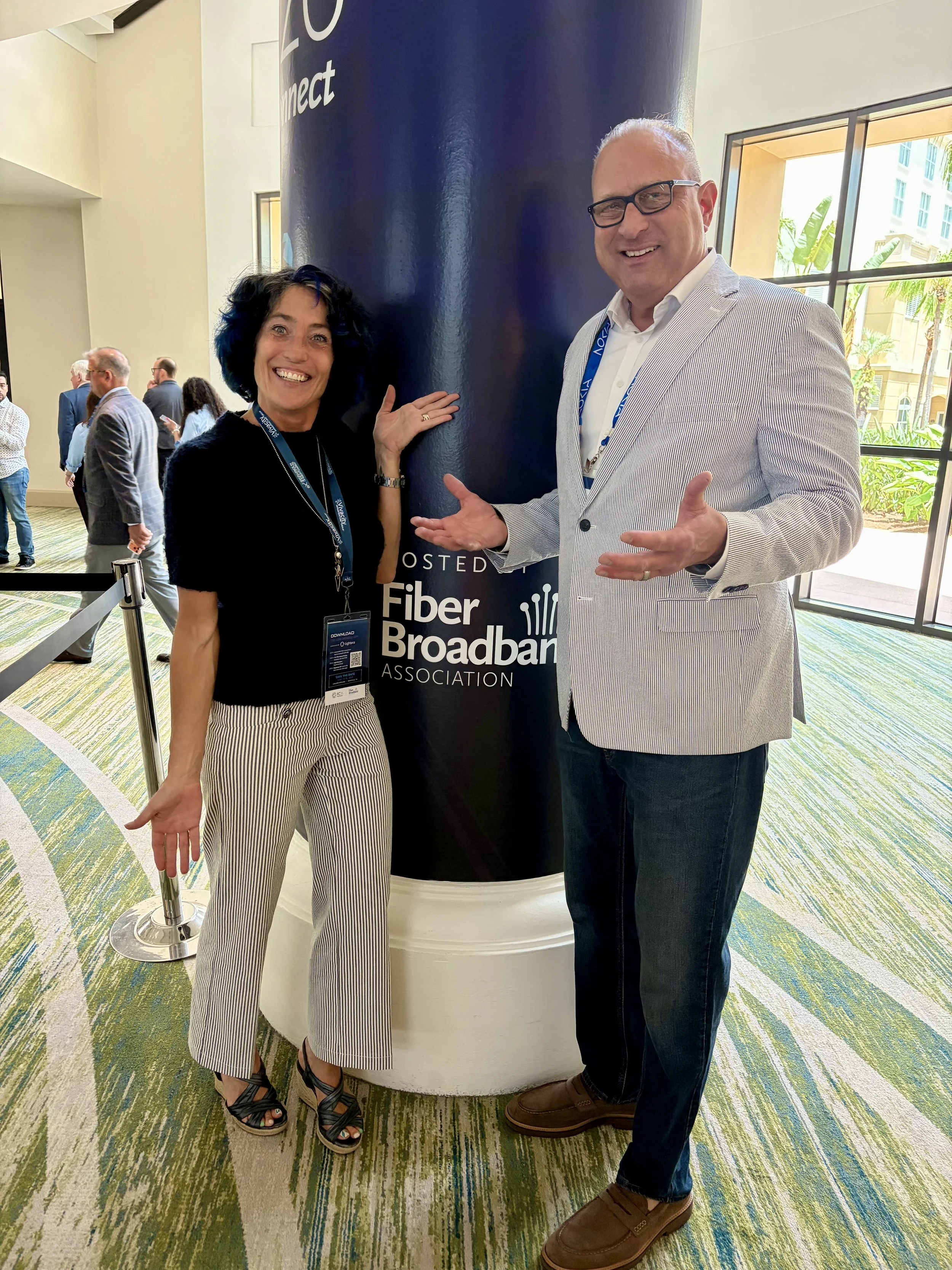 Two people smiling and gesturing towards a vertical pole with the words 'Fiber Broadband' on it. They are at a conference or event, wearing name tags and standing in a room with large windows and other attendees in the background.