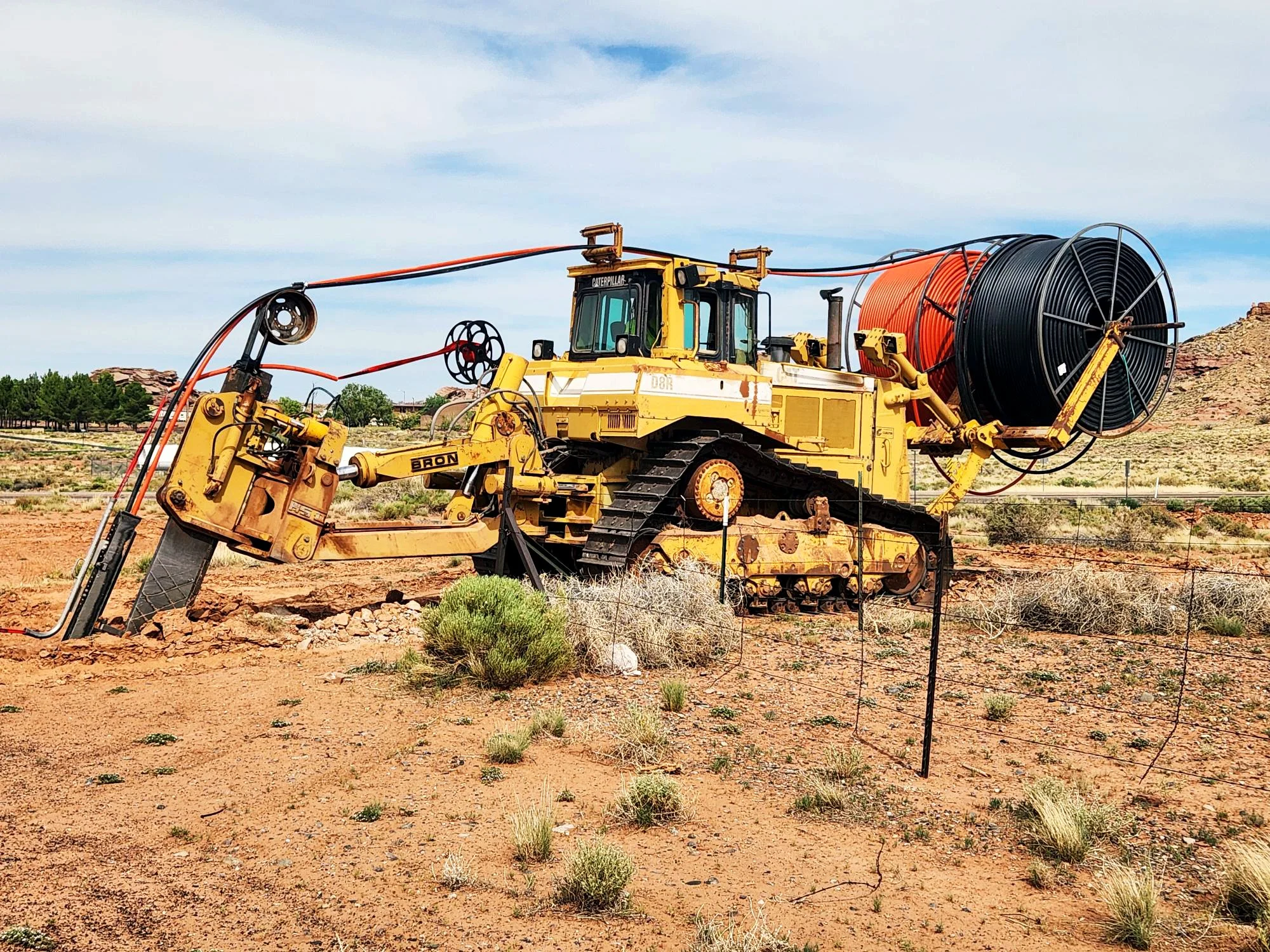 A large piece of construction equipment with two spools of cable, one black and one orange, situated in a dry, arid landscape with sparse vegetation and a distant rocky hill under a cloudy sky.