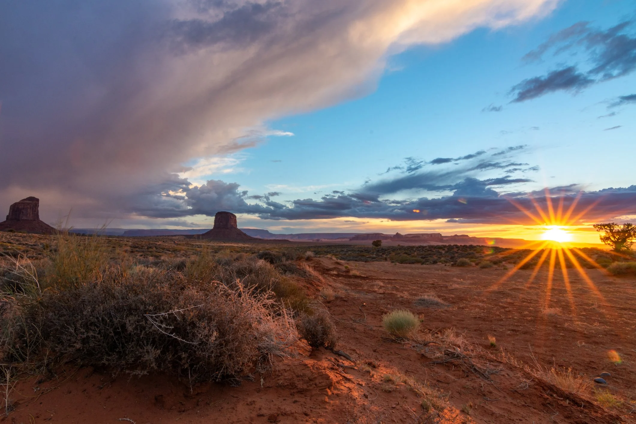 Sunset over a desert landscape with large rock formations and scattered bushes in the foreground. The sky has dark clouds on the left and blue sky with some clouds on the right, with the sun setting on the horizon creating a bright flare.