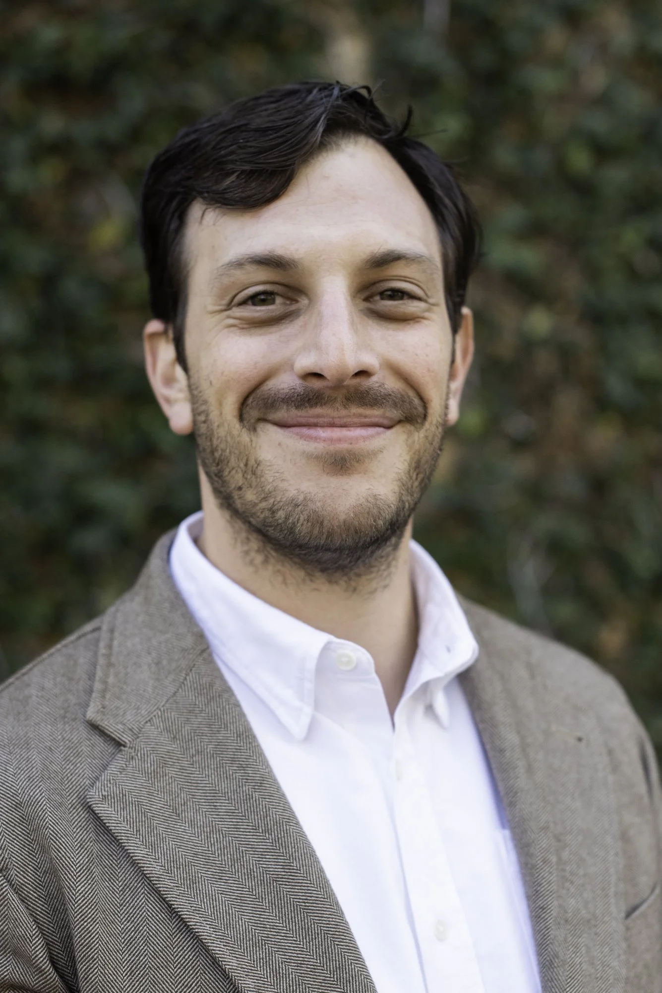 Close-up of a man smiling outdoors, wearing a white shirt and a brown tweed blazer, with a blurred green natural background.