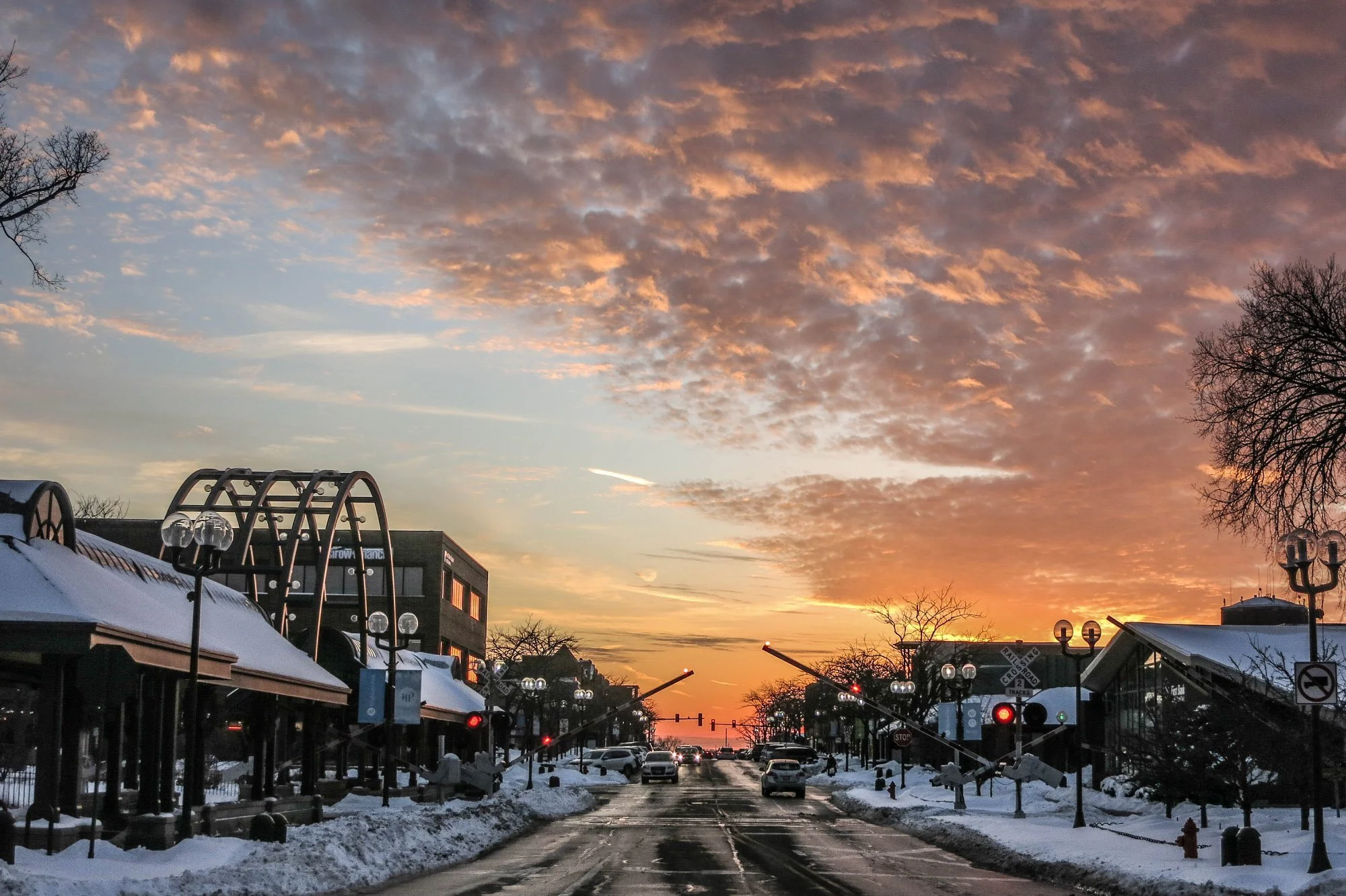 A city street at sunset with shops on either side, snow-covered sidewalks, lampposts, cars on the road, and trees with bare branches under an orange and pink cloud-filled sky.