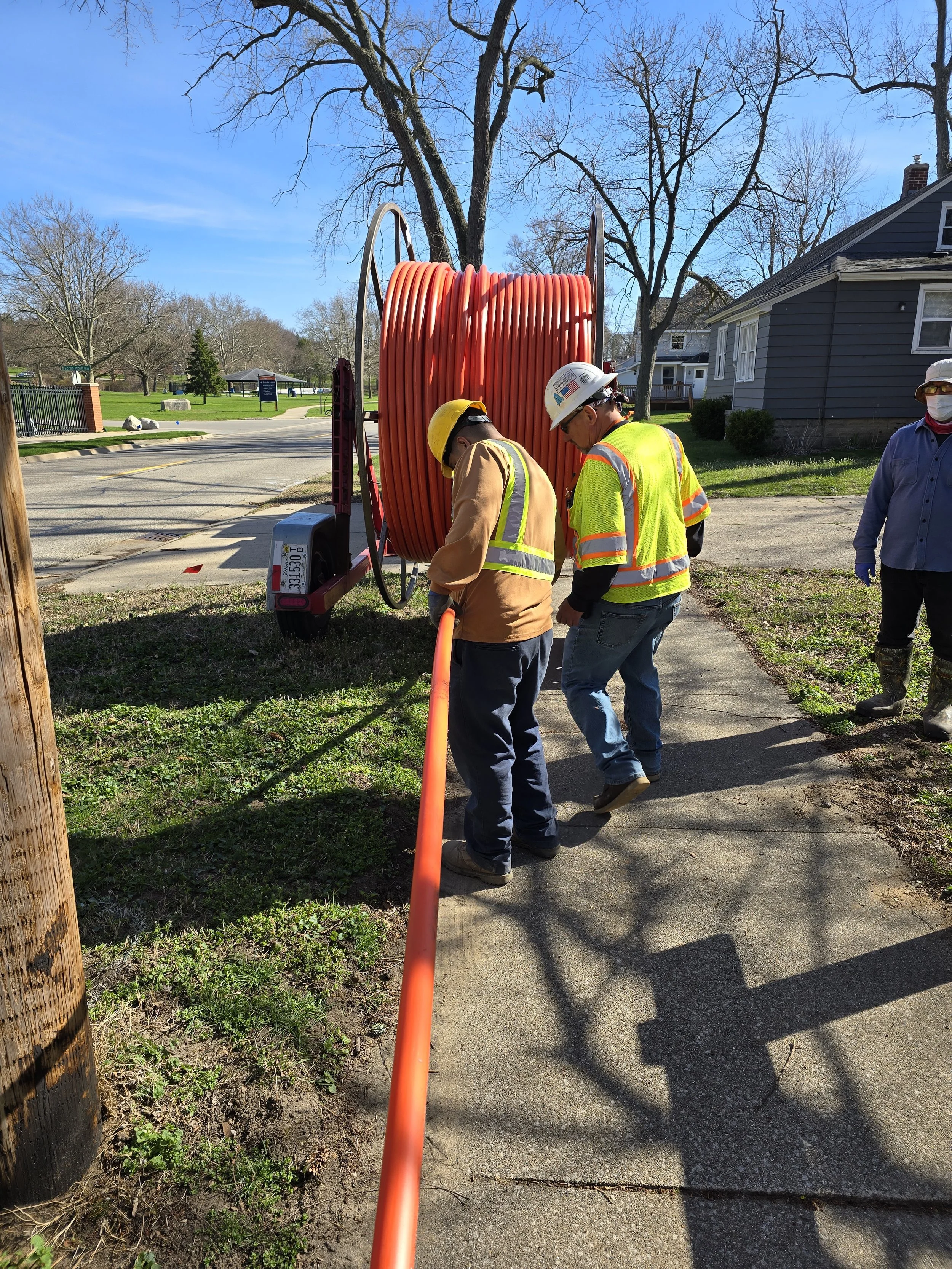 Two construction workers wearing safety vests and helmets working near an orange utility line reel on a sidewalk in a suburban neighborhood.