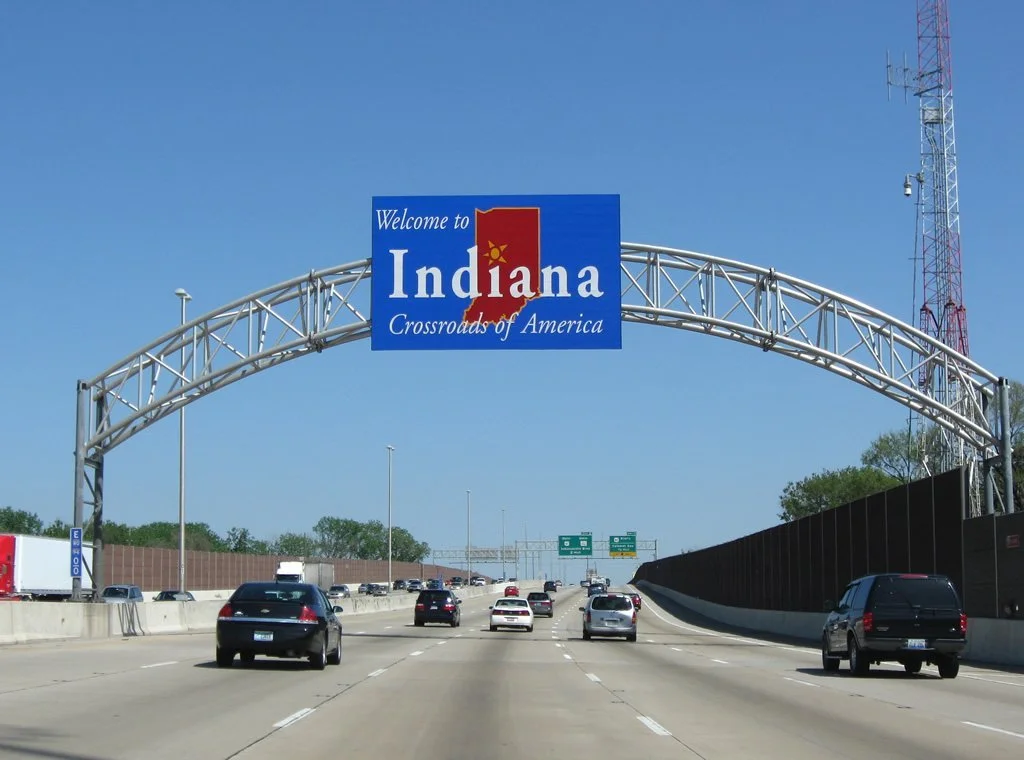 Highway with cars and a large blue welcome sign that reads 'Welcome to Indiana, Crossroads of America' with a red letter I and a yellow star, under a clear blue sky.