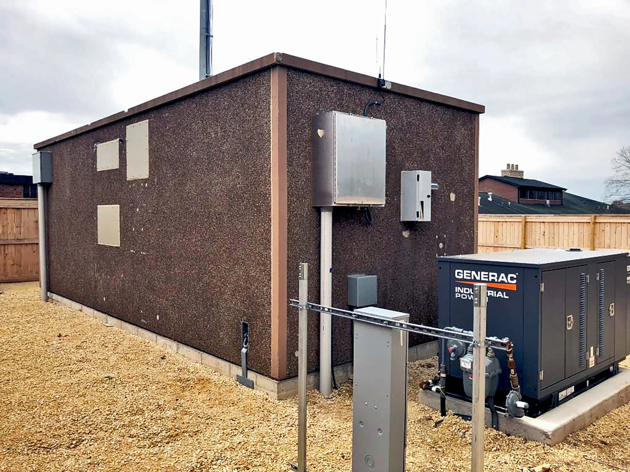 Outdoor view of a brown building with electrical equipment, including a generator and panels, under an cloudy sky.
