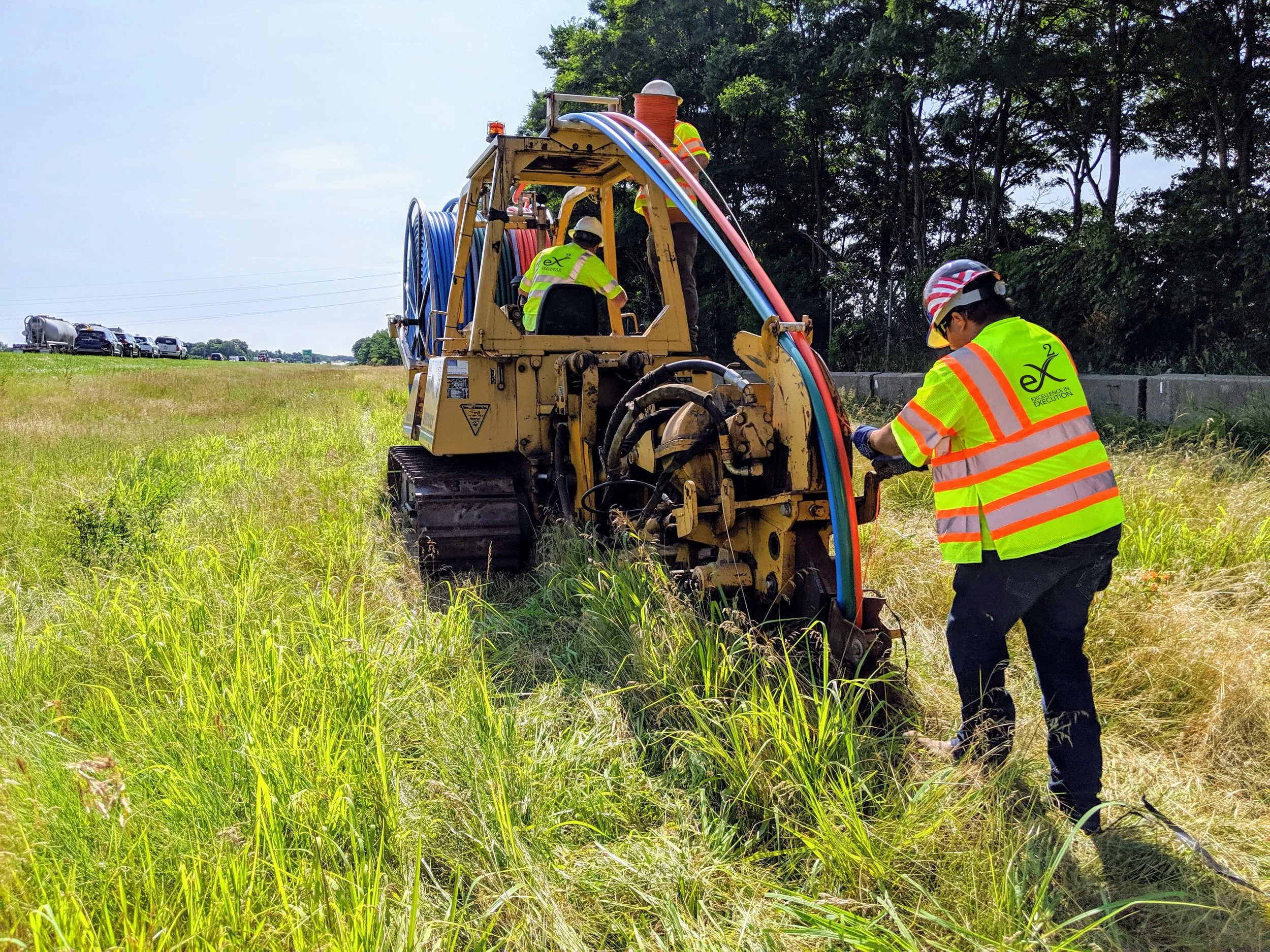 Workers in bright yellow safety vests and helmets operate a small construction or maintenance machine with pipes, near a grassy field and a line of vehicles on a road, with trees in the background.