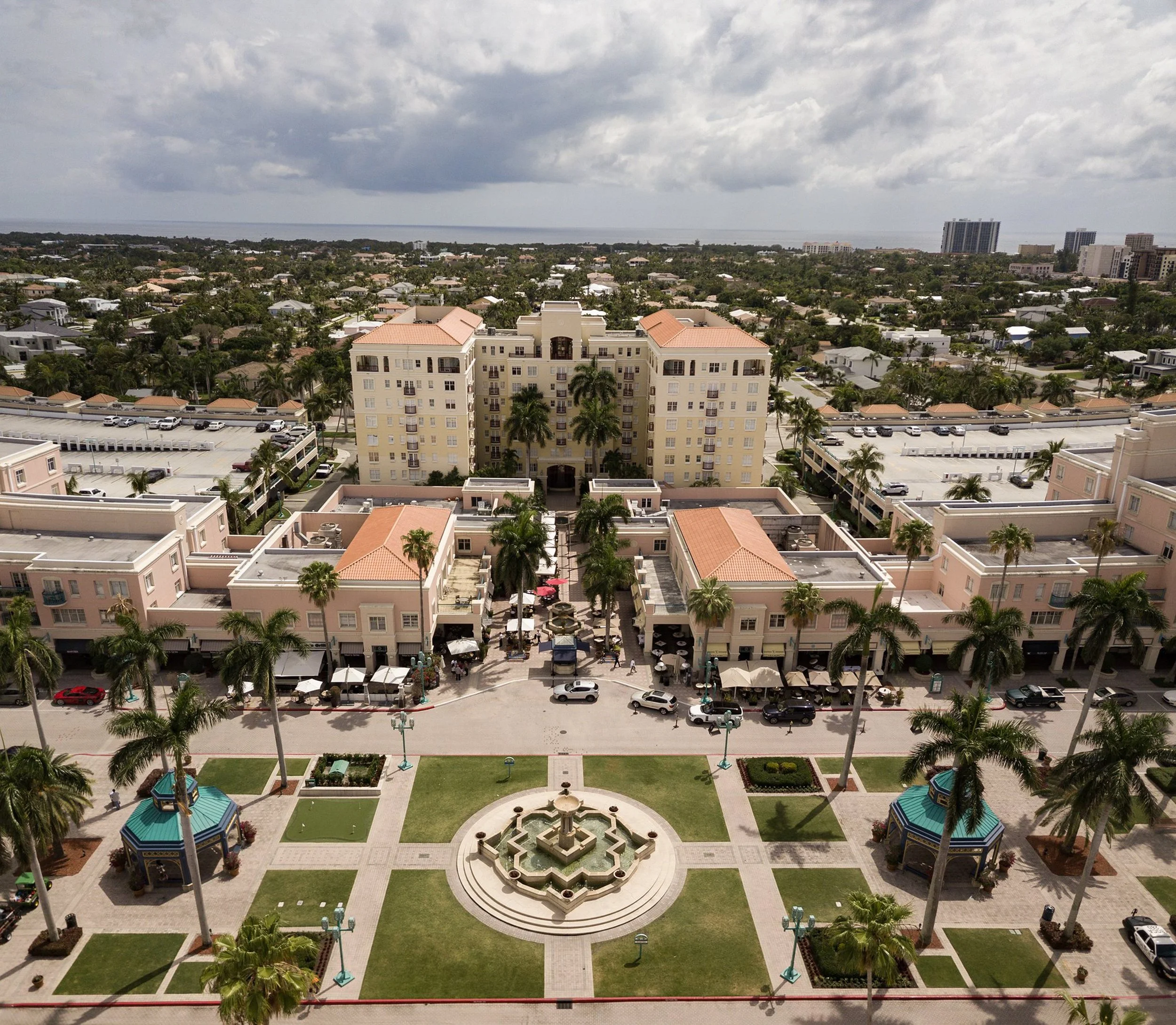 Aerial view of a city with palm trees, a fountain, and a large beige apartment building with orange roofs. The cityscape extends to the ocean in the distance, with tall buildings and cloudy skies overhead.