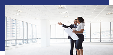Two professionals, a man and a woman, discussing and pointing towards an empty conference room with large windows and city skyline views.
