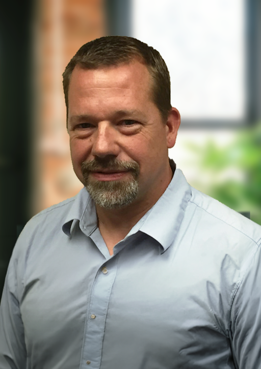 A middle-aged man with short brown hair, a beard, and a mustache wearing a light blue collared shirt, standing indoors with a blurred background of a window and greenery.