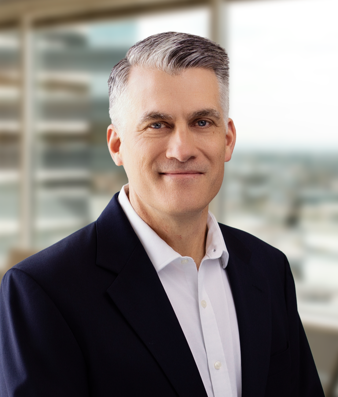 A middle-aged man with short gray hair wearing a black suit and white shirt, smiling in an office setting with large windows in the background.