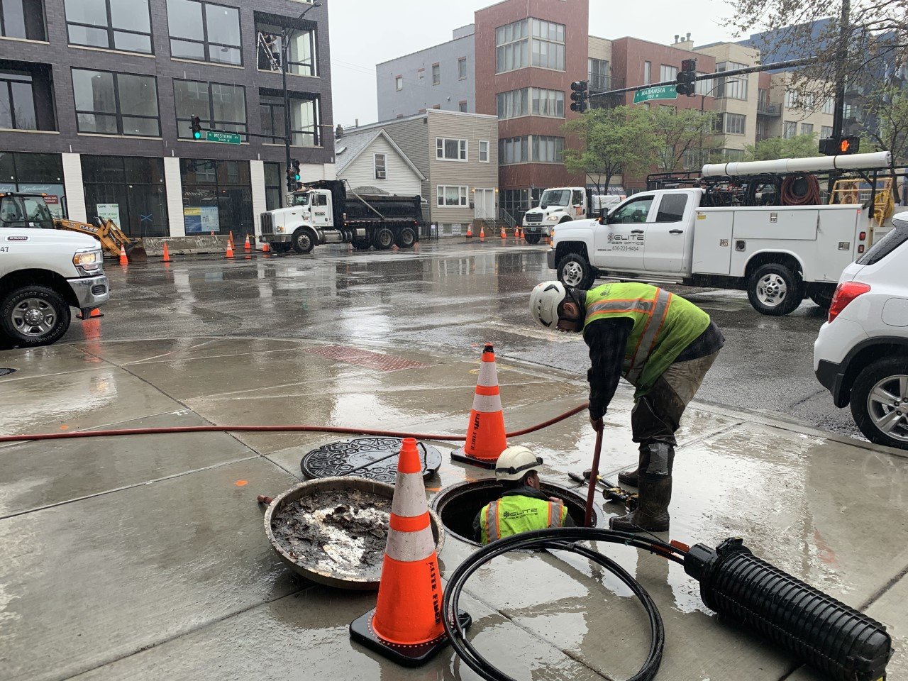 Construction workers repairing a street drainage system on a rainy day, with orange safety cones and construction vehicles nearby.