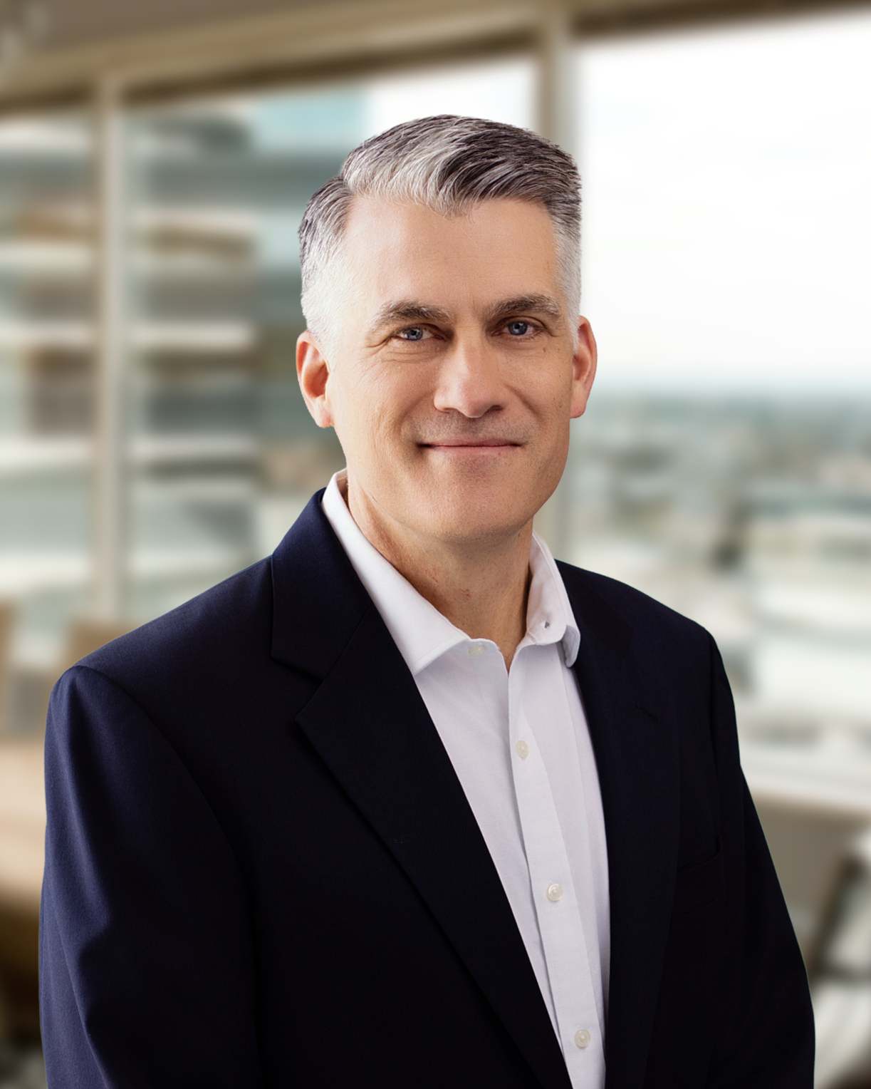Professional man with gray hair, wearing a dark suit and white dress shirt, standing in a modern office building with large windows and blurred cityscape background.