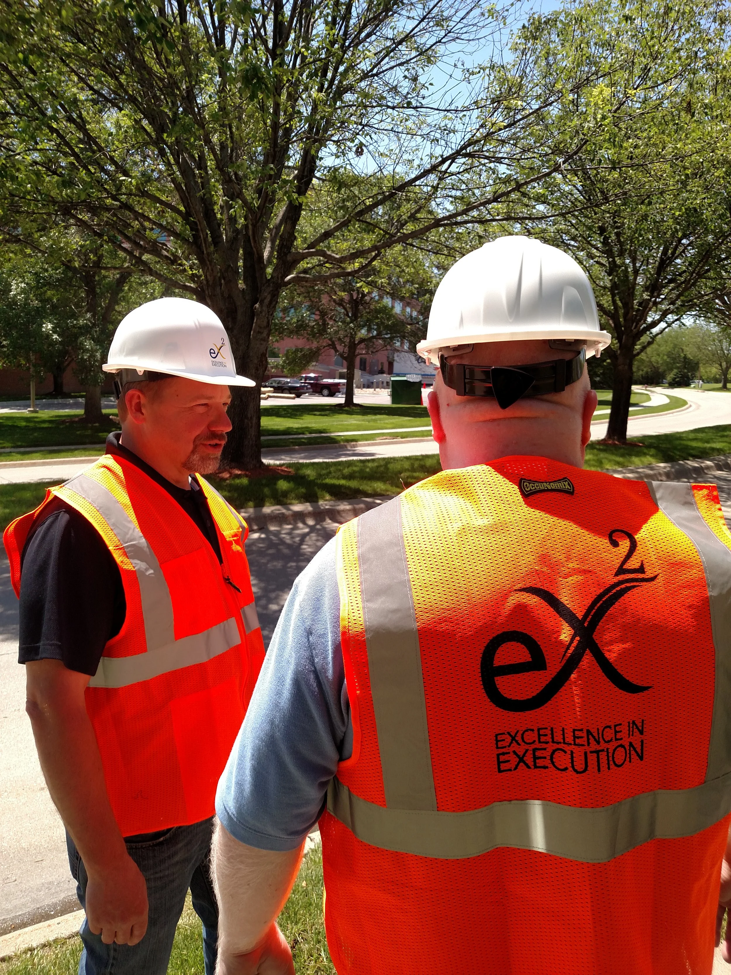 Two men wearing hard hats and safety vests standing outdoors in a park-like area with trees and a building in the background. One man's vest has the phrase 'Excellence in Execution' on it.