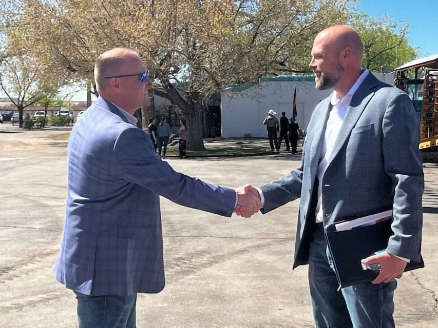 Two men in business suits shaking hands outdoors on a sunny day in front of a tree and some people standing near a building.