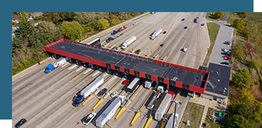Aerial view of a toll plaza with multiple lanes and a red covered overhead structure, traffic moving through the lanes, and cars waiting in line to pay tolls.