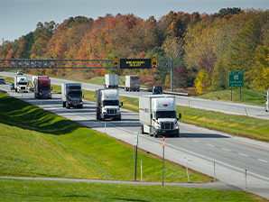 Multiple semi-trucks driving on a highway surrounded by colorful fall foliage