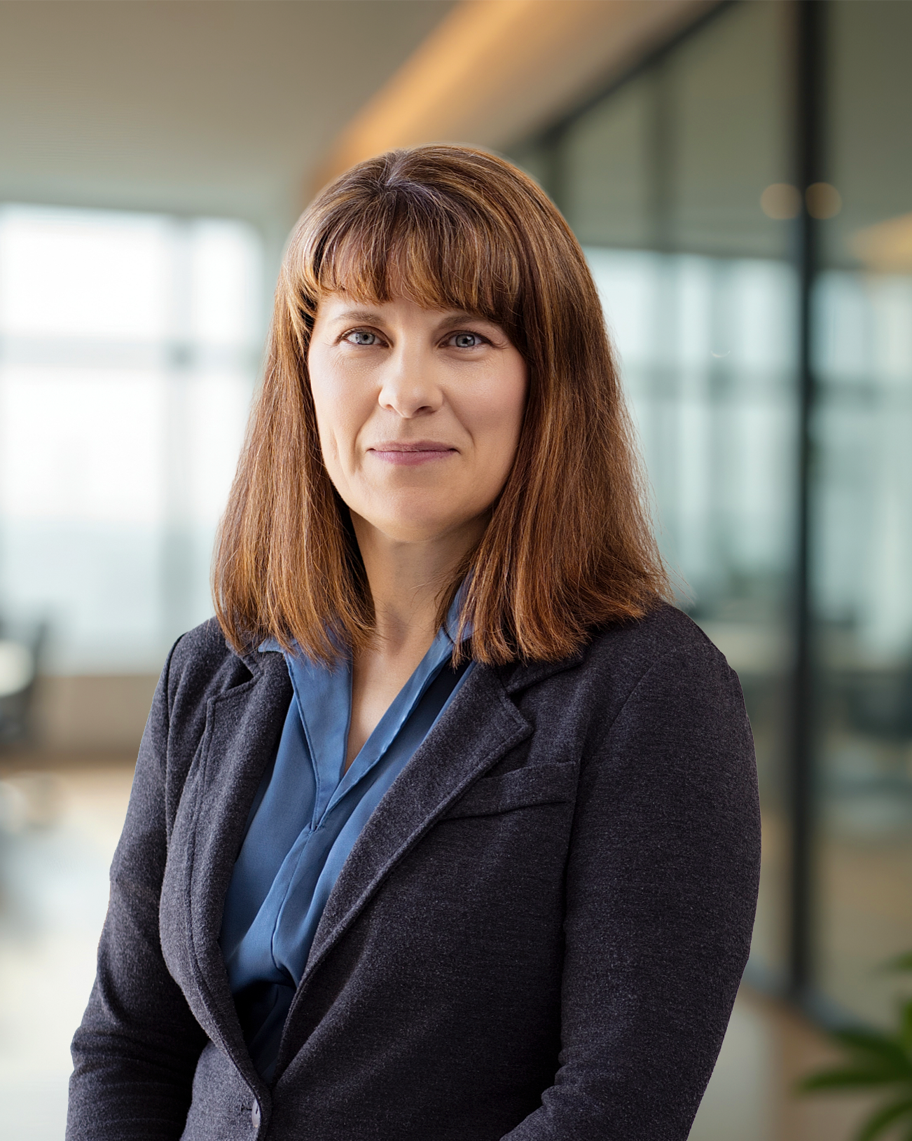 A professional woman with brown hair and blue eyes wearing a blue shirt and dark blazer, standing in a modern office environment.
