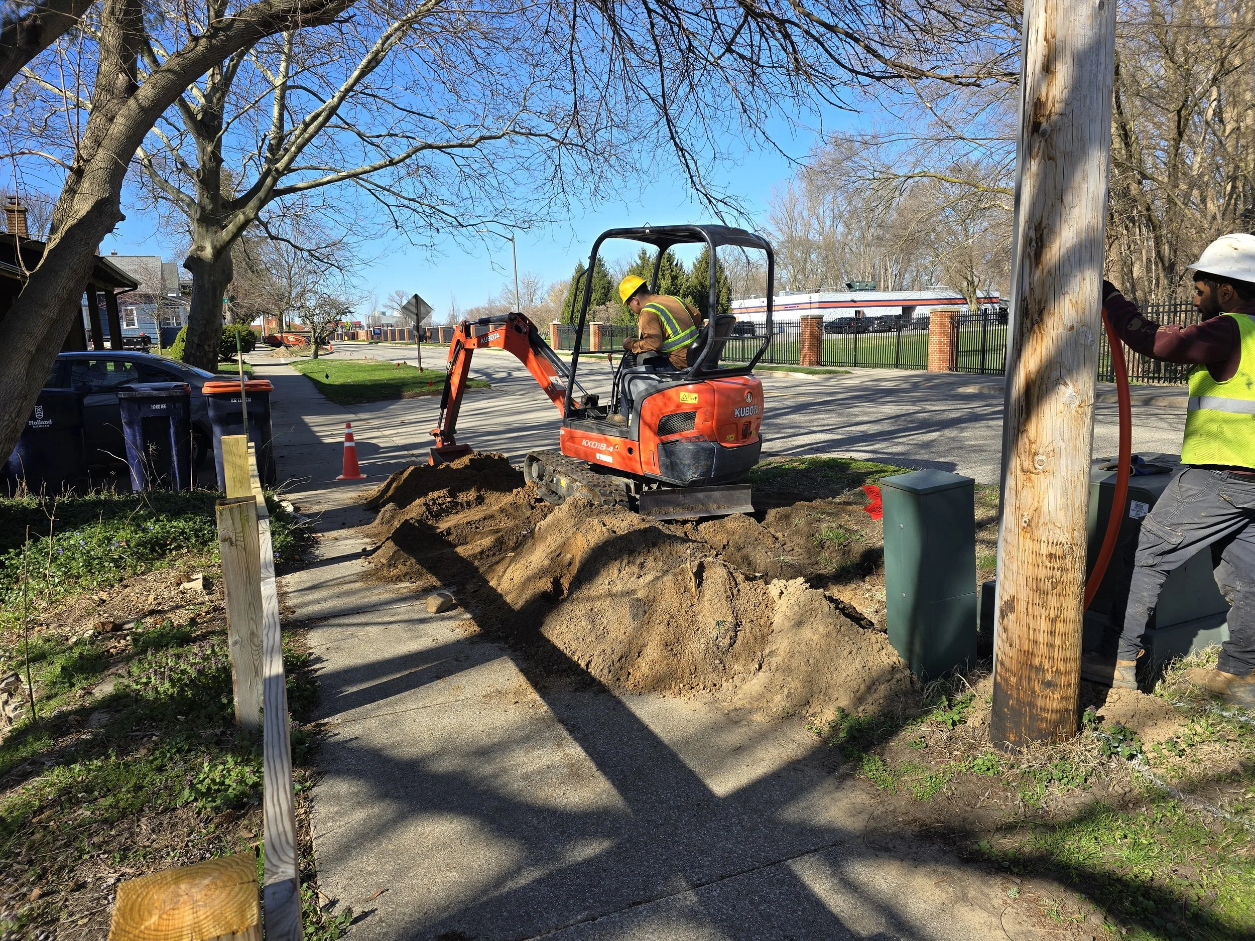 Construction workers excavating dirt on a sidewalk near a utility pole, with trees, cars, and buildings in the background.