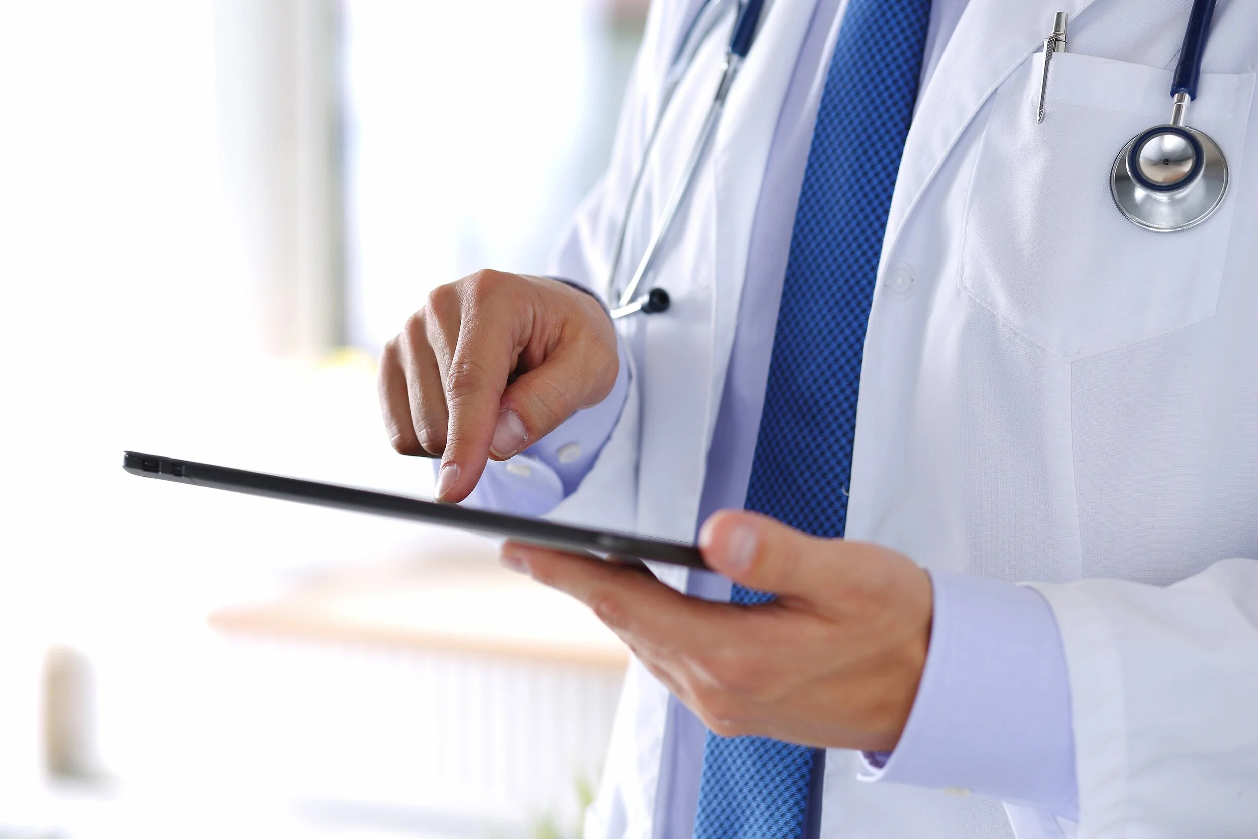 A healthcare professional wearing a white coat with a stethoscope around their neck, holding a tablet device.