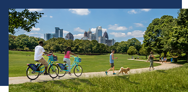 People walking and biking with a dog in a park, skyline in the background.