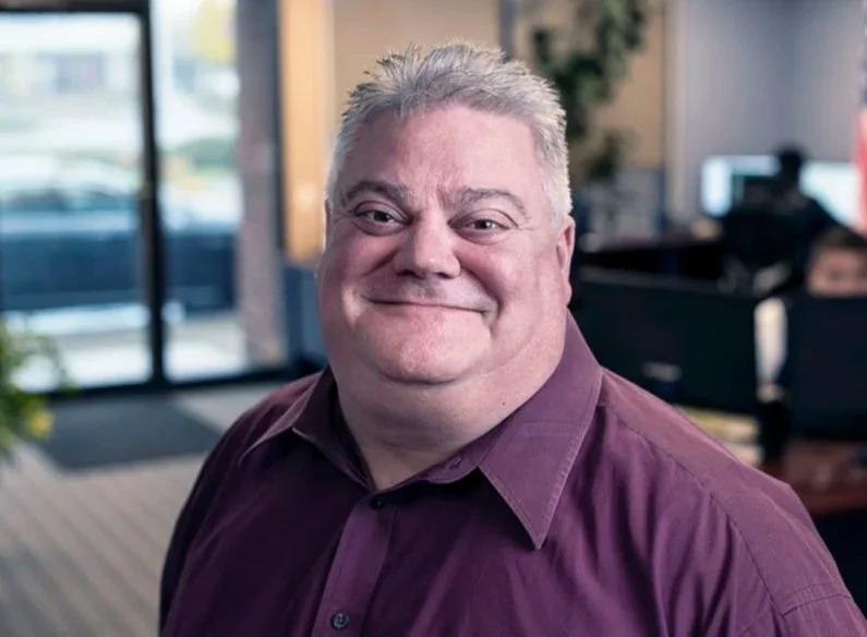 Smiling middle-aged man with short gray hair, wearing a purple dress shirt, in an office setting with people working at desks in the background.