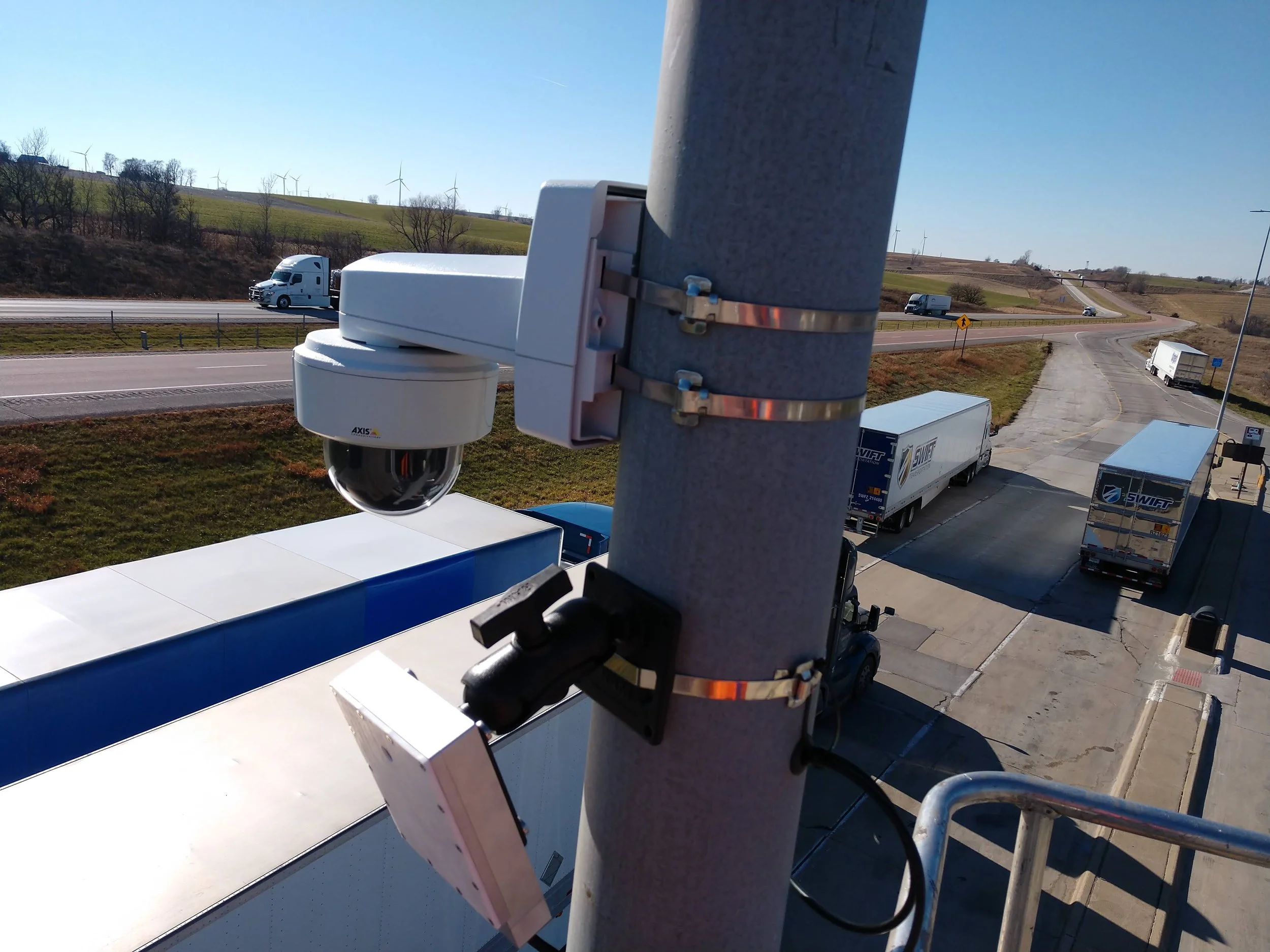 A security camera mounted on a metal pole overlooking a highway with trucks traveling on it and wind turbines on hills in the background.