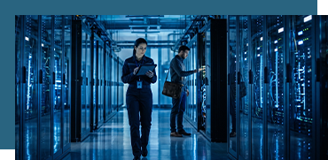 Two IT professionals working in a server room with blue lighting, one holding a tablet, the other inspecting server equipment.
