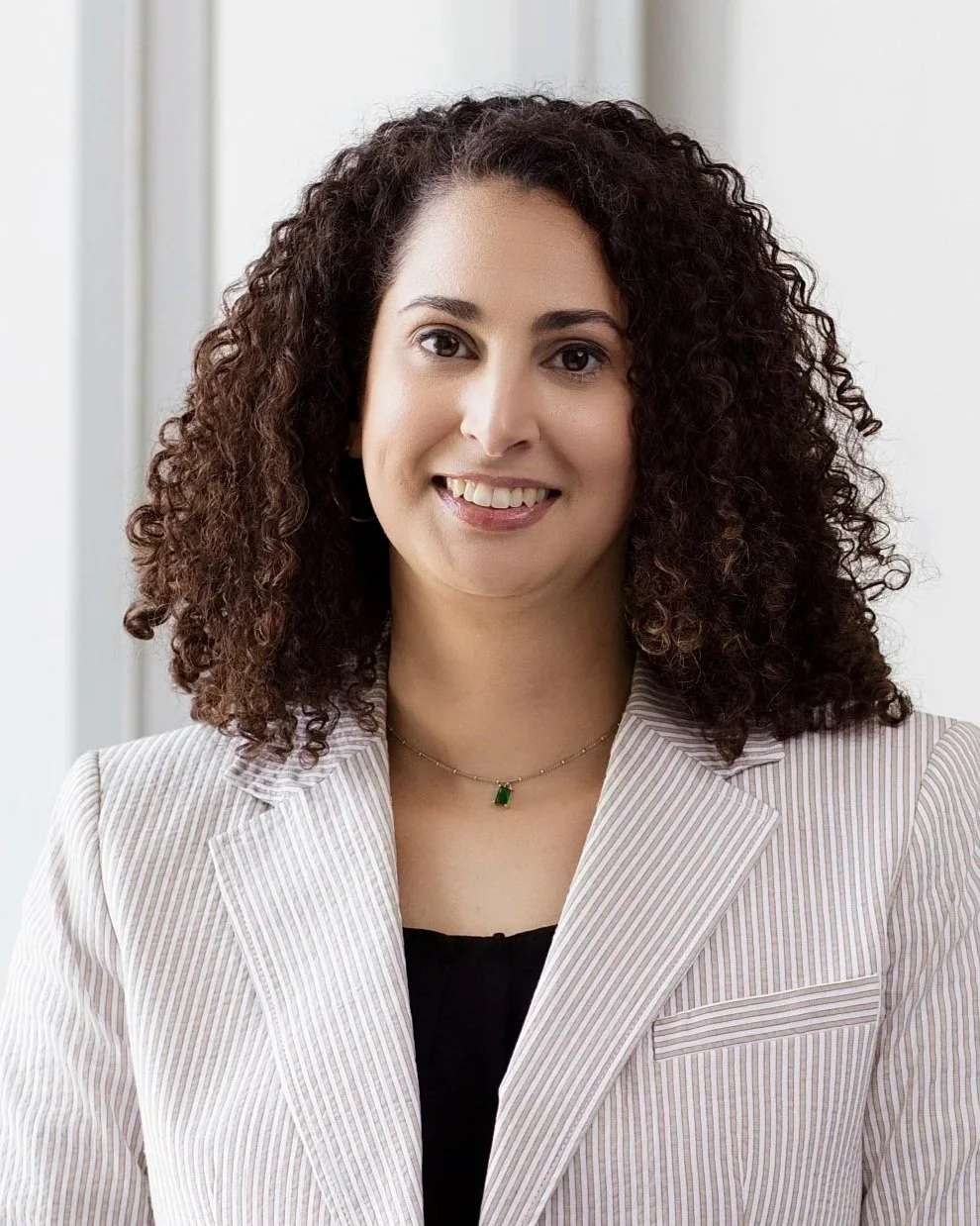 A professional woman with curly dark hair, wearing a light-colored striped blazer and a necklace with a green pendant, smiling indoors near a window.
