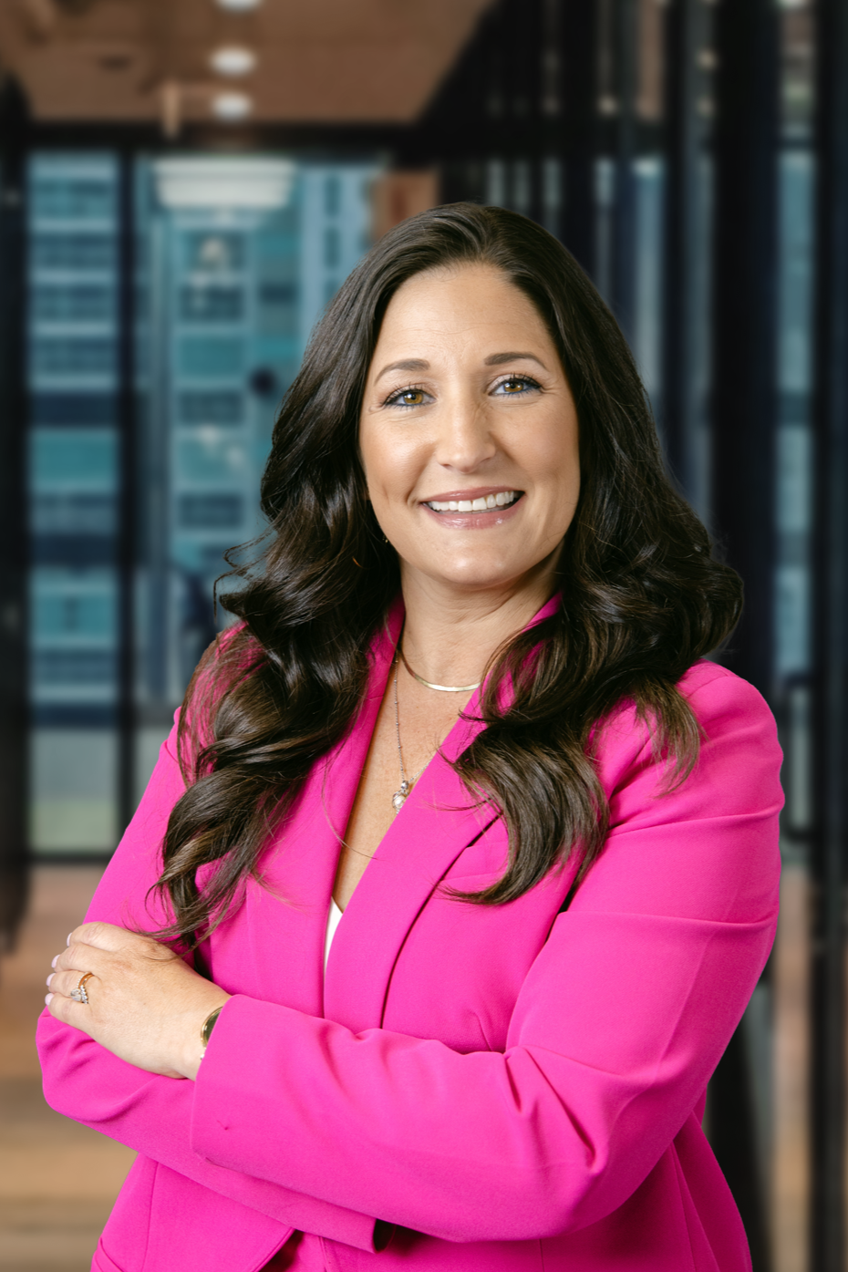 A smiling professional woman with long dark wavy hair, wearing a bright pink blazer, standing in a modern office with large windows and city buildings in the background.