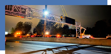 Nighttime scene of a highway with emergency vehicles, flashing lights, trees, and a large overhead structure.