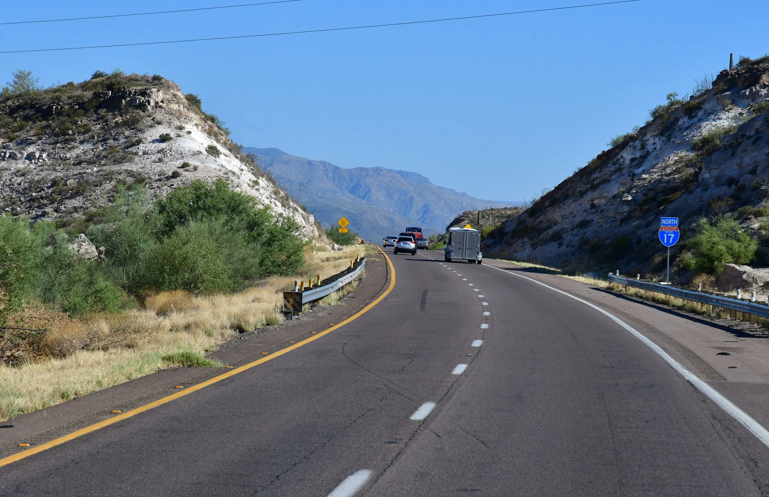 A two-lane highway running through a mountainous desert landscape with rocky hills on both sides and a clear blue sky. Several cars and a trailer are visible on the road. A North Interstate 17 road sign is on the right side.