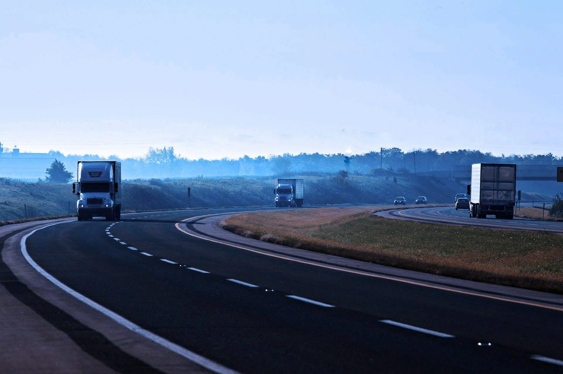 A highway with multiple trucks and cars traveling on it, surrounded by green grassy areas and rolling hills under a blue sky with some clouds.