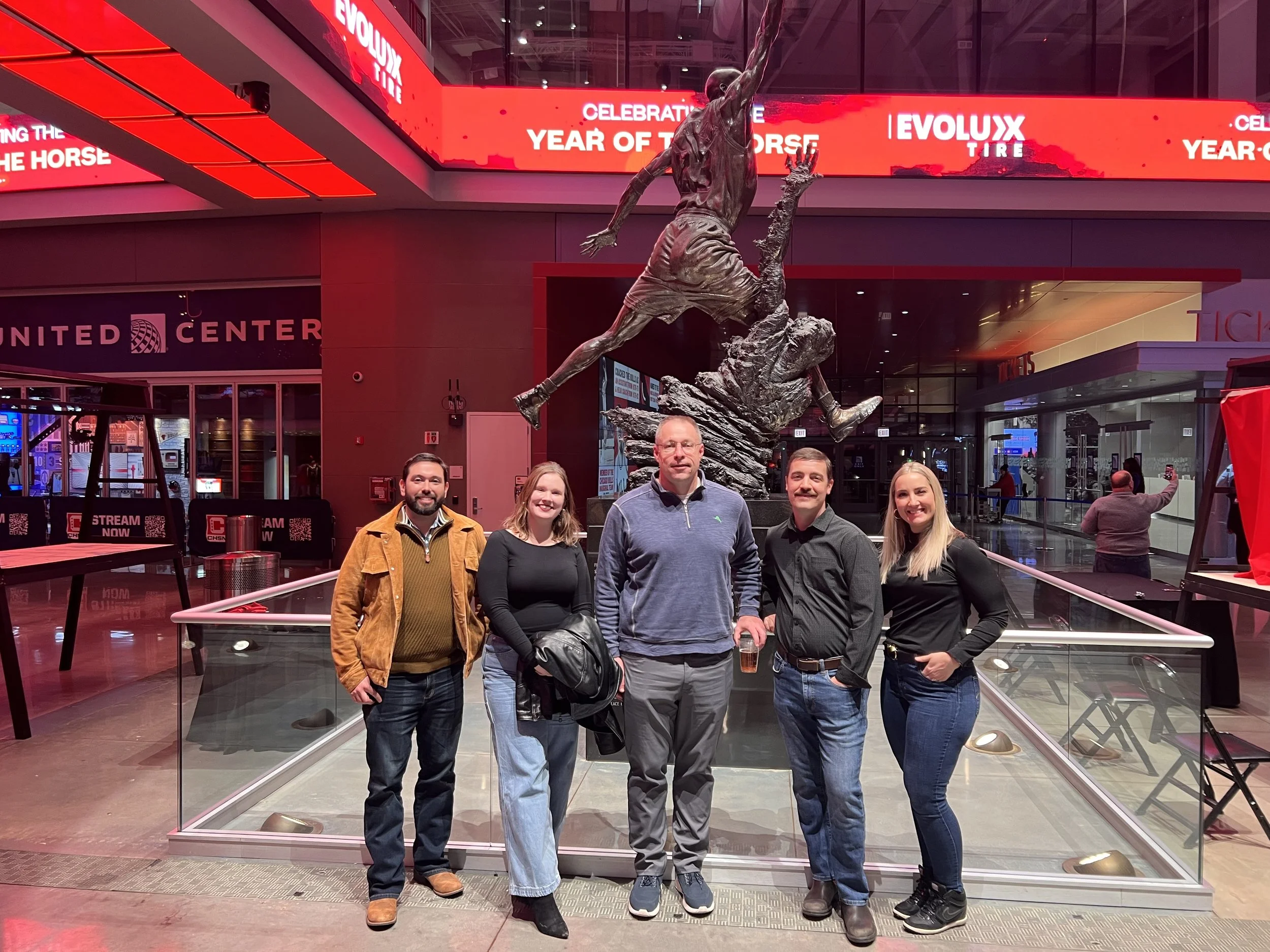 Five people standing inside a glass enclosure in a building at night. A large statue of a running athlete is in the background, and red digital signs are visible above and behind the group.