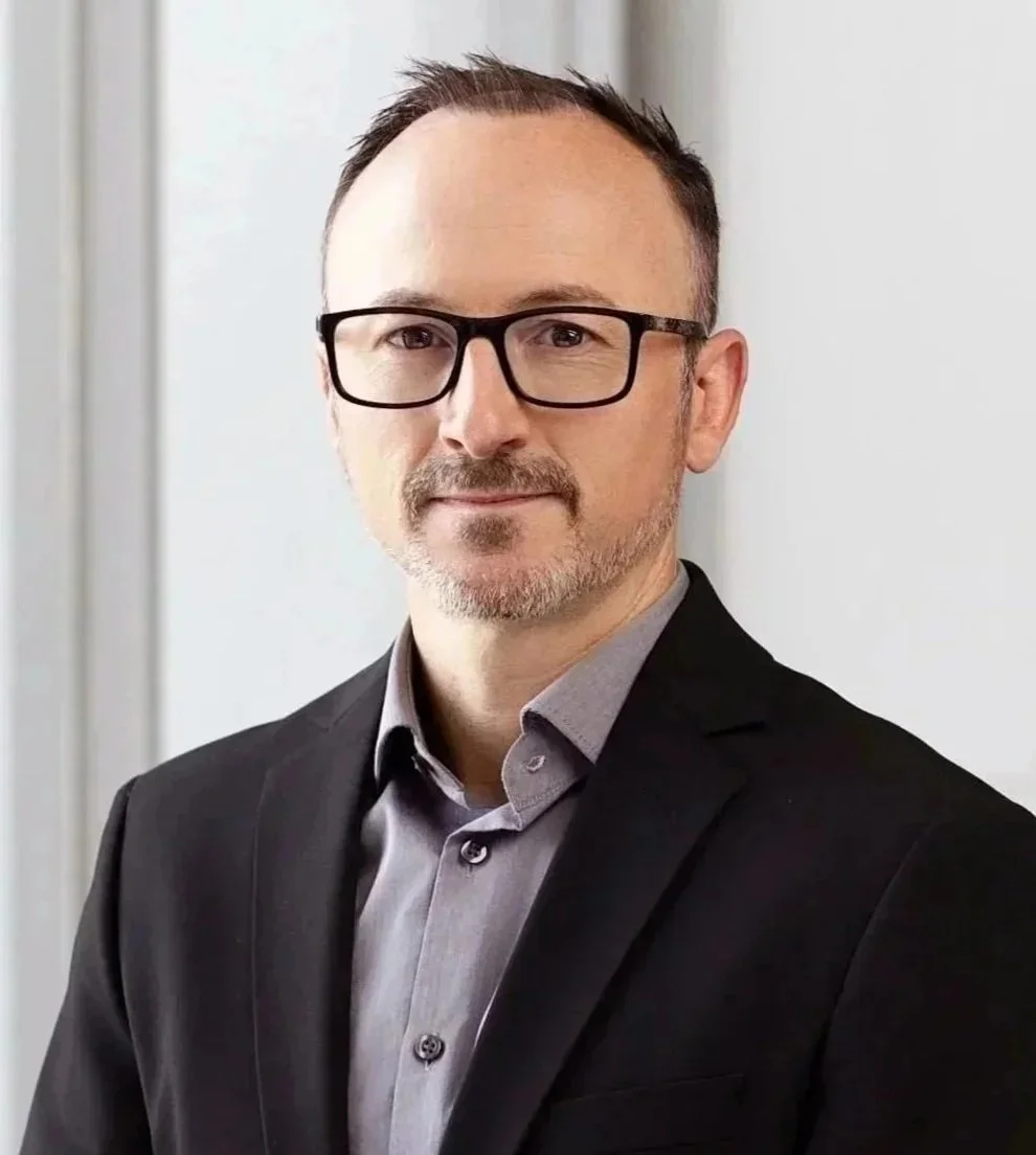 A man with glasses, short dark hair, and a beard, wearing a suit and standing indoors against a plain background.