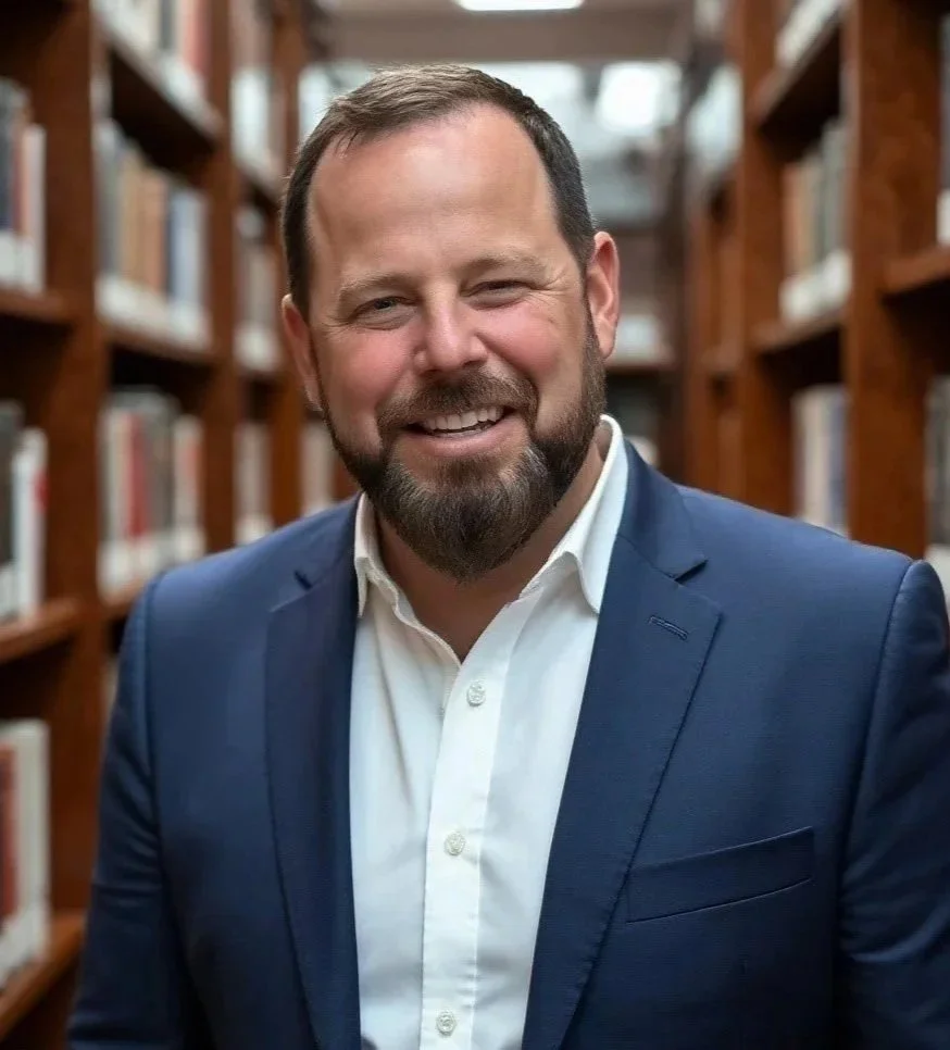 Smiling man in a navy suit and white shirt standing in a library with wooden shelves of books in the background.