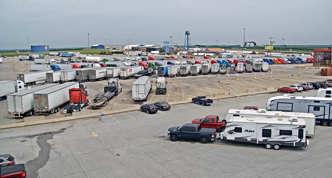 An airport parking lot filled with semi-trucks, cars, and RVs, with an airport terminal and water tower in the background.