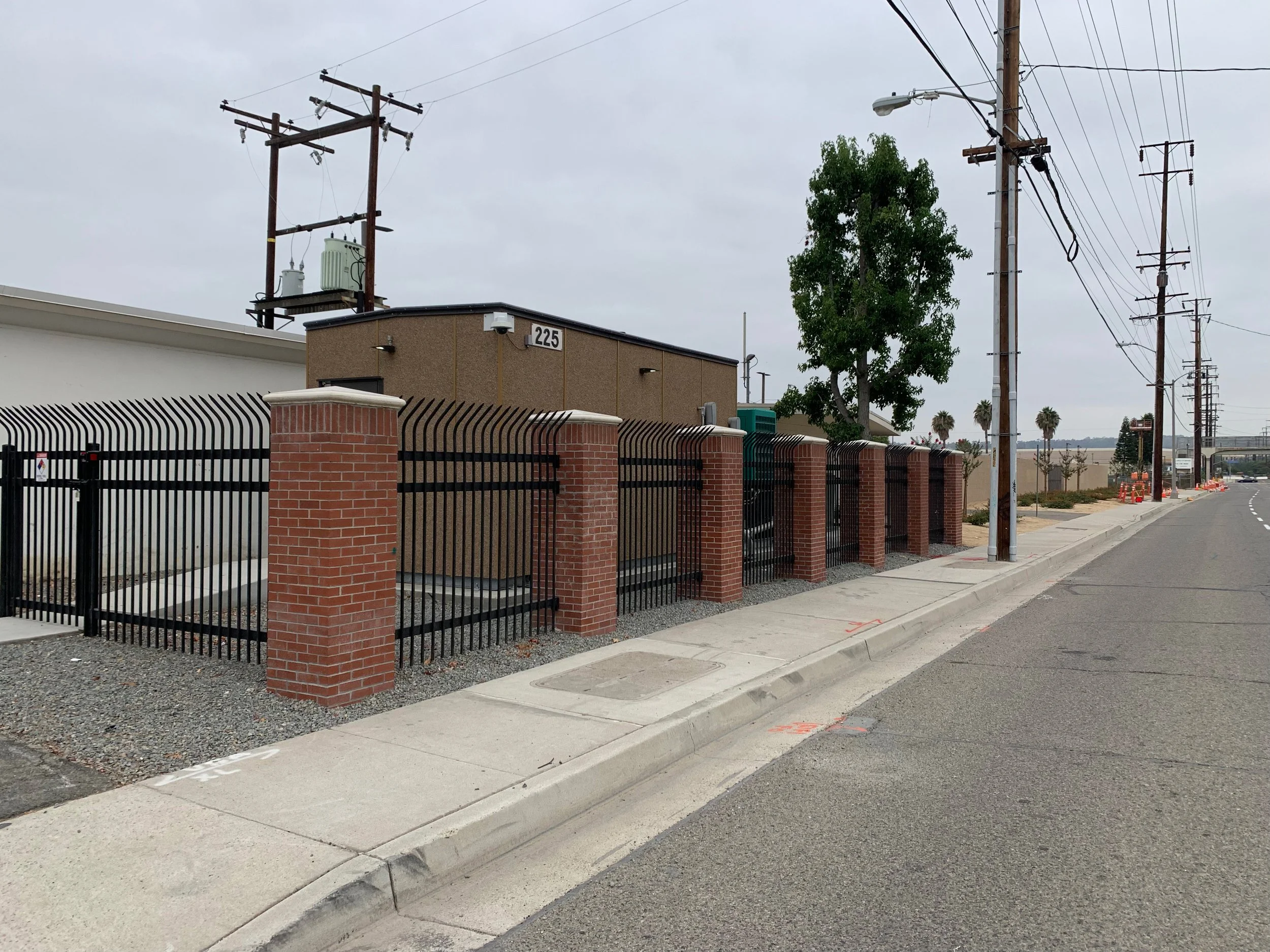 Street view with utility poles, a brick fence, a brown building marked 225, and a tree under an overcast sky.