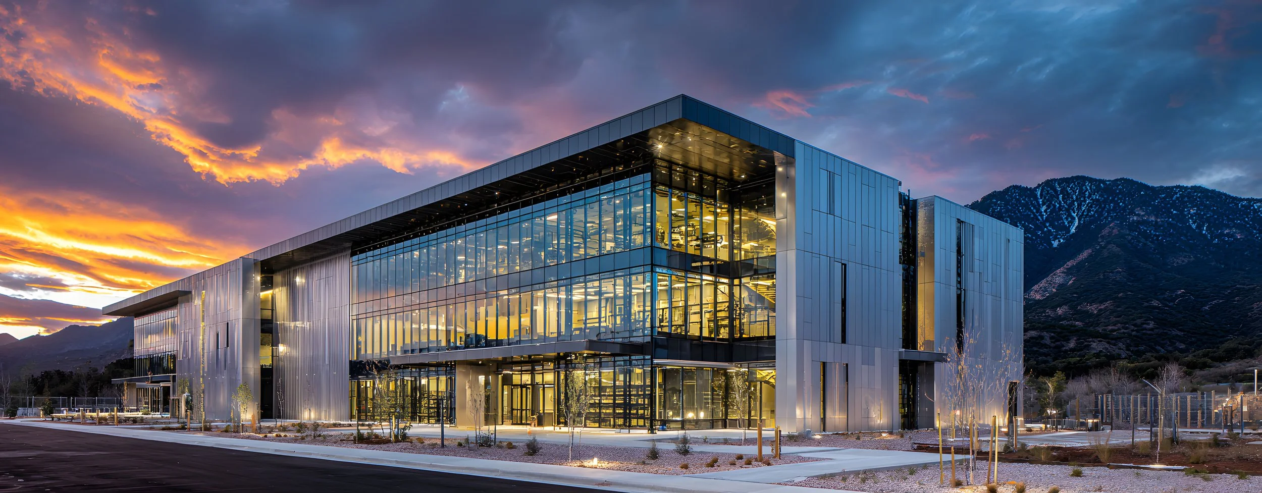 Modern glass building with illuminated interior, mountains in background, cloudy sunset sky