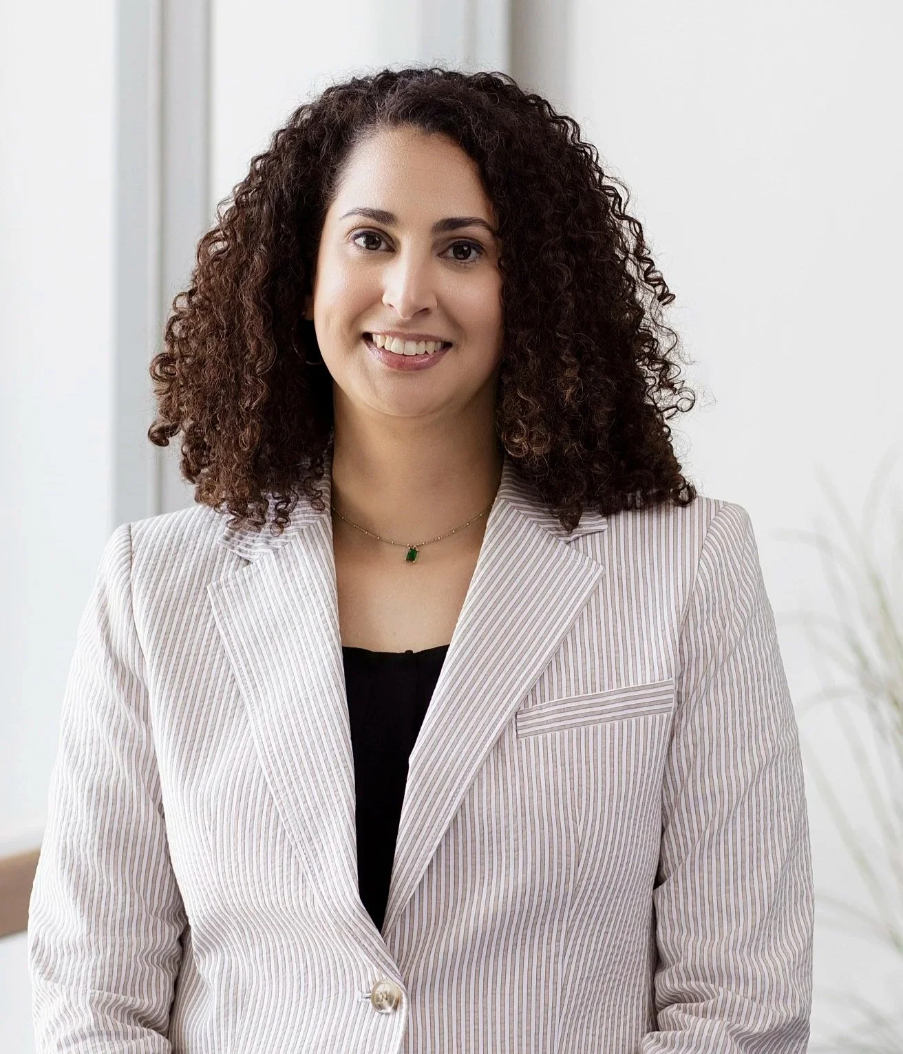 A woman with curly dark hair smiling, wearing a striped blazer and a green pendant necklace, standing indoors near a window.