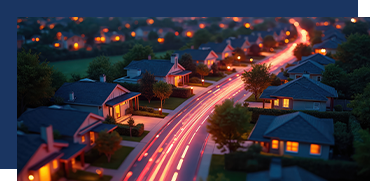 Nighttime suburban neighborhood with illuminated houses and a curving street with car light trails.