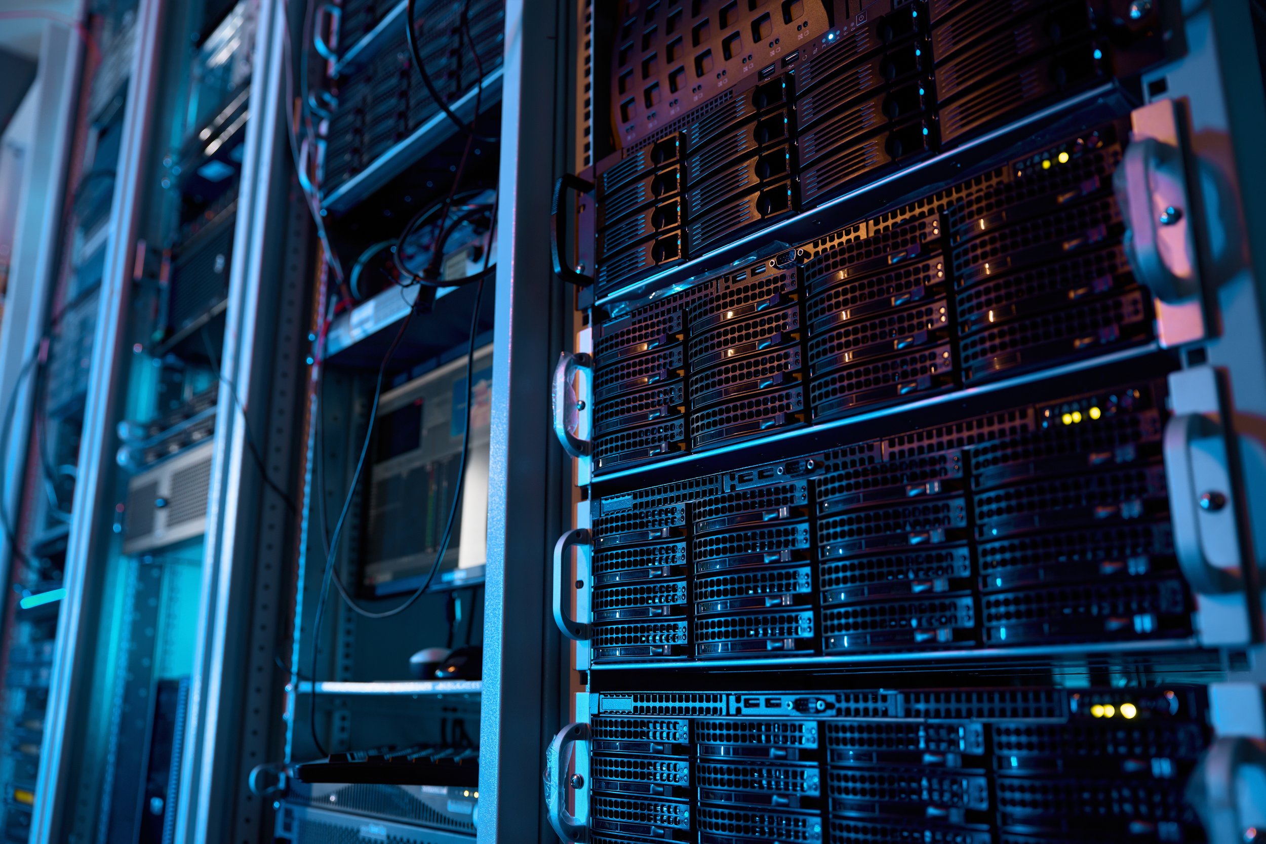 A server room with multiple black server racks filled with hardware components, illuminated by blue and warm orange lights.
